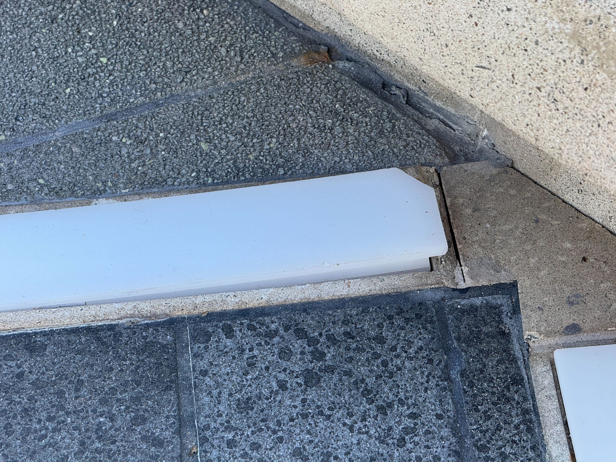 Close-up of a stair edge with chipped and misaligned tiles and a white stair nosing strip.
