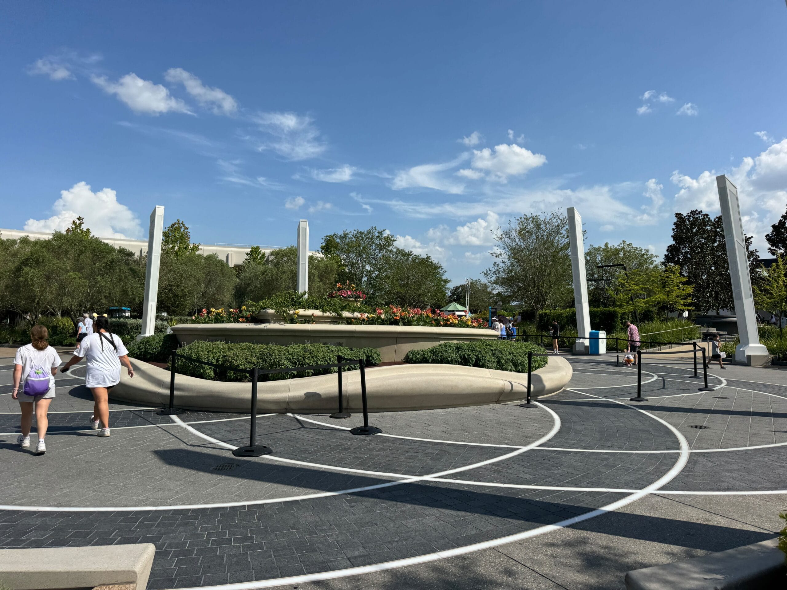 People walk near a circular flower bed and tall pillars in a landscaped public area under a blue sky.
