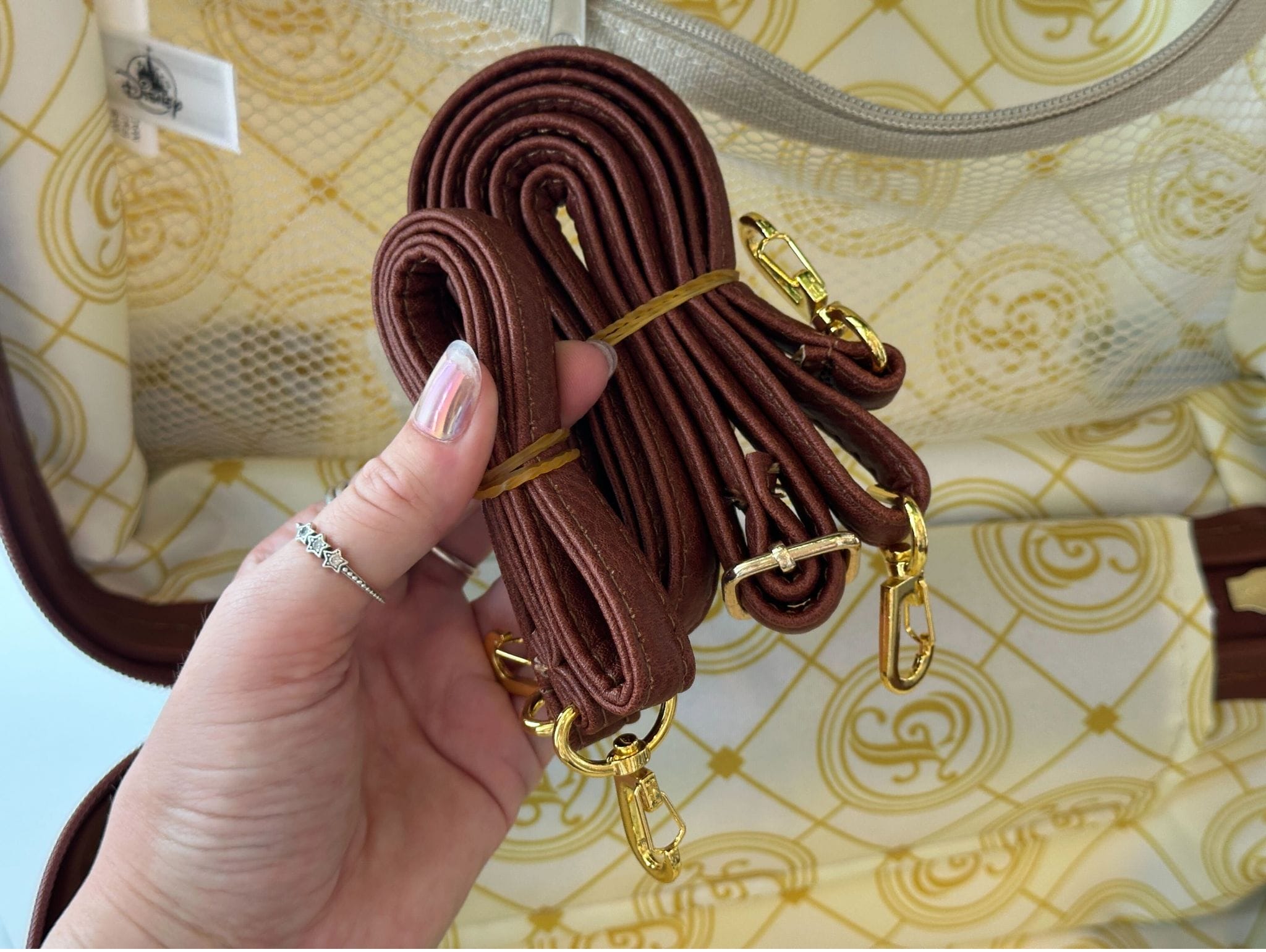 A hand holds two coiled brown bag straps with gold clasps over a Disney resort mini luggage bag’s patterned beige interior.