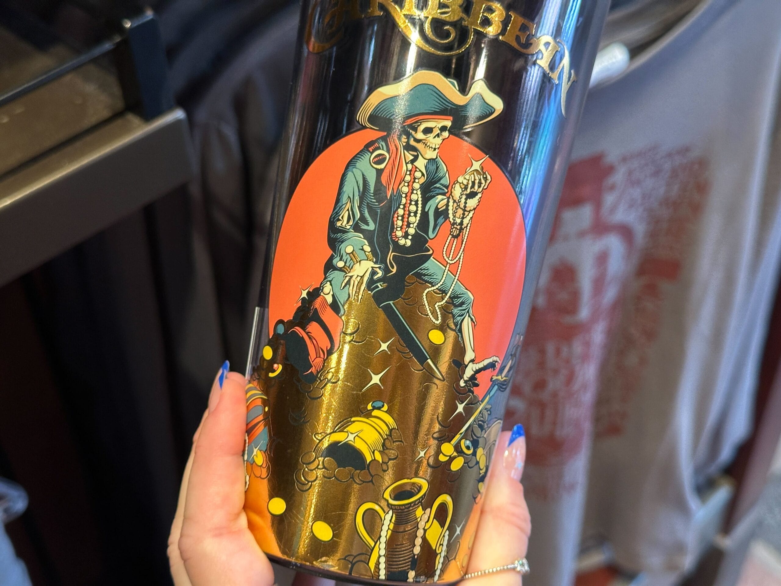 A hand holds a tumbler featuring a Pirates of the Caribbean-style skeleton pirate with sword, gold coins, and treasure chests.