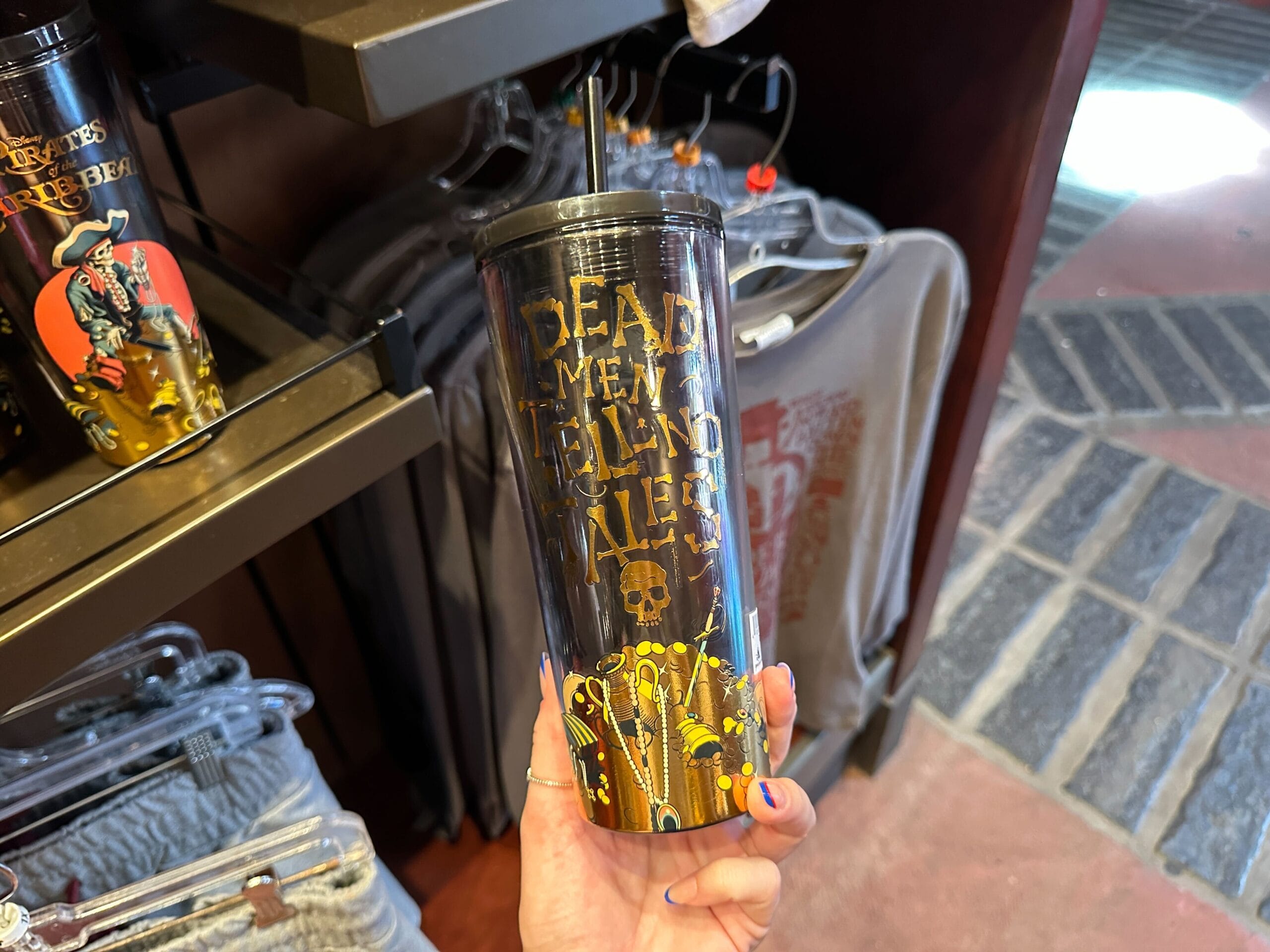 A hand holds a black Pirates of the Caribbean tumbler with gold "Dead men tell no tales" text in a retail store.