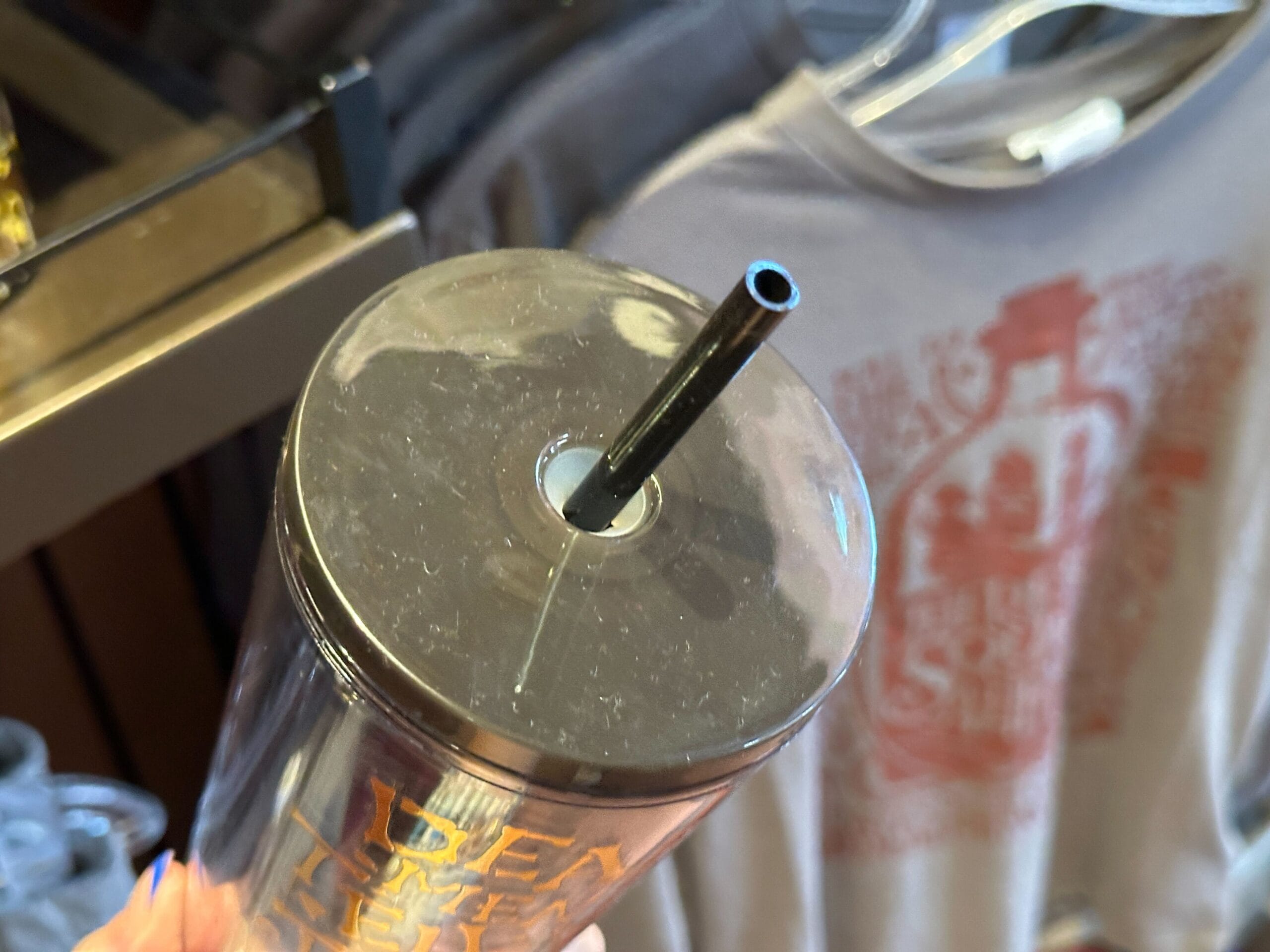 A close-up of a tumbler lid with a black straw, held before a Pirates of the Caribbean graphic tee on a rack.