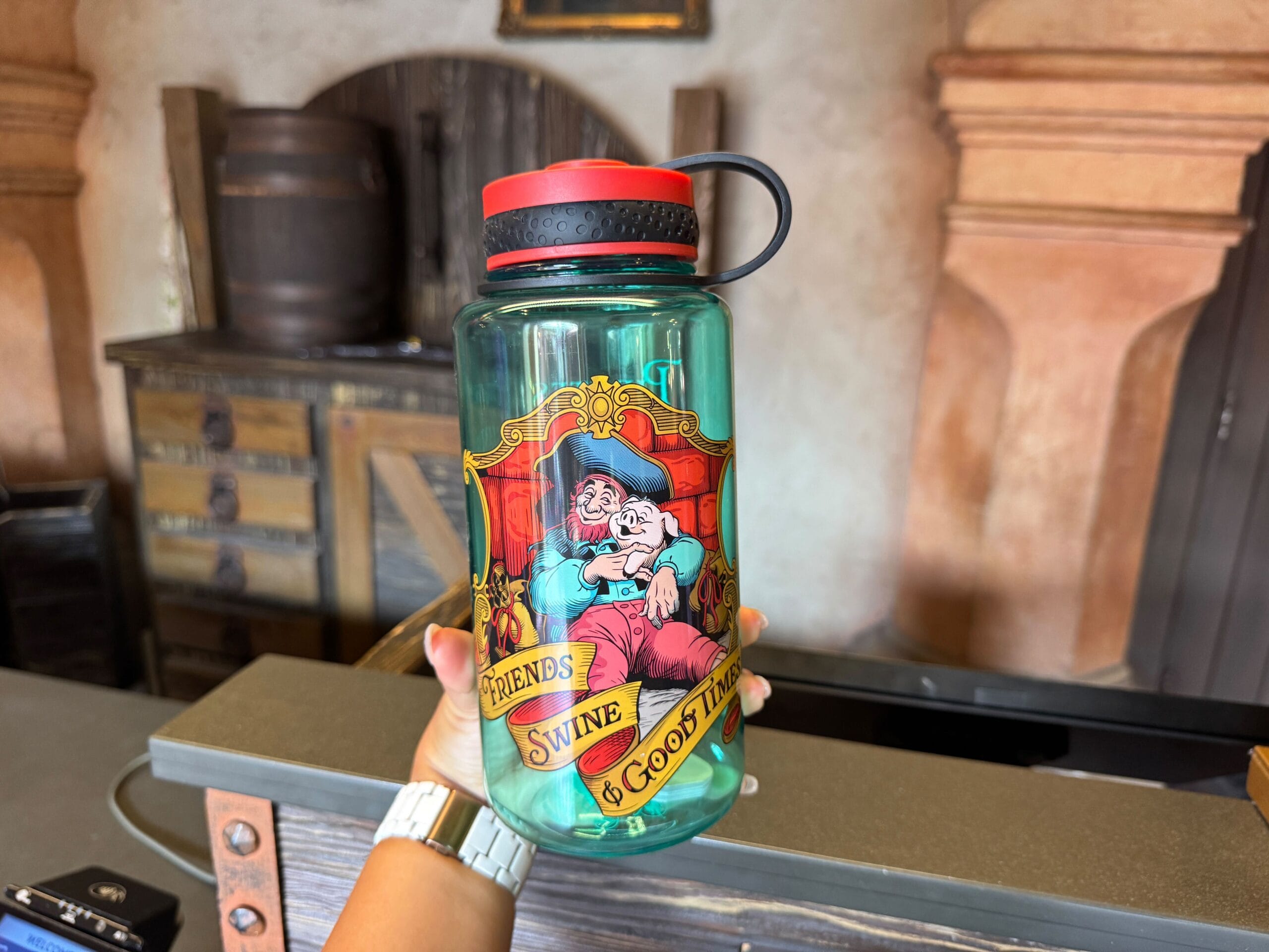 A hand holds a water bottle featuring cartoon pigs with the text "Friends, Swine & Good Times.