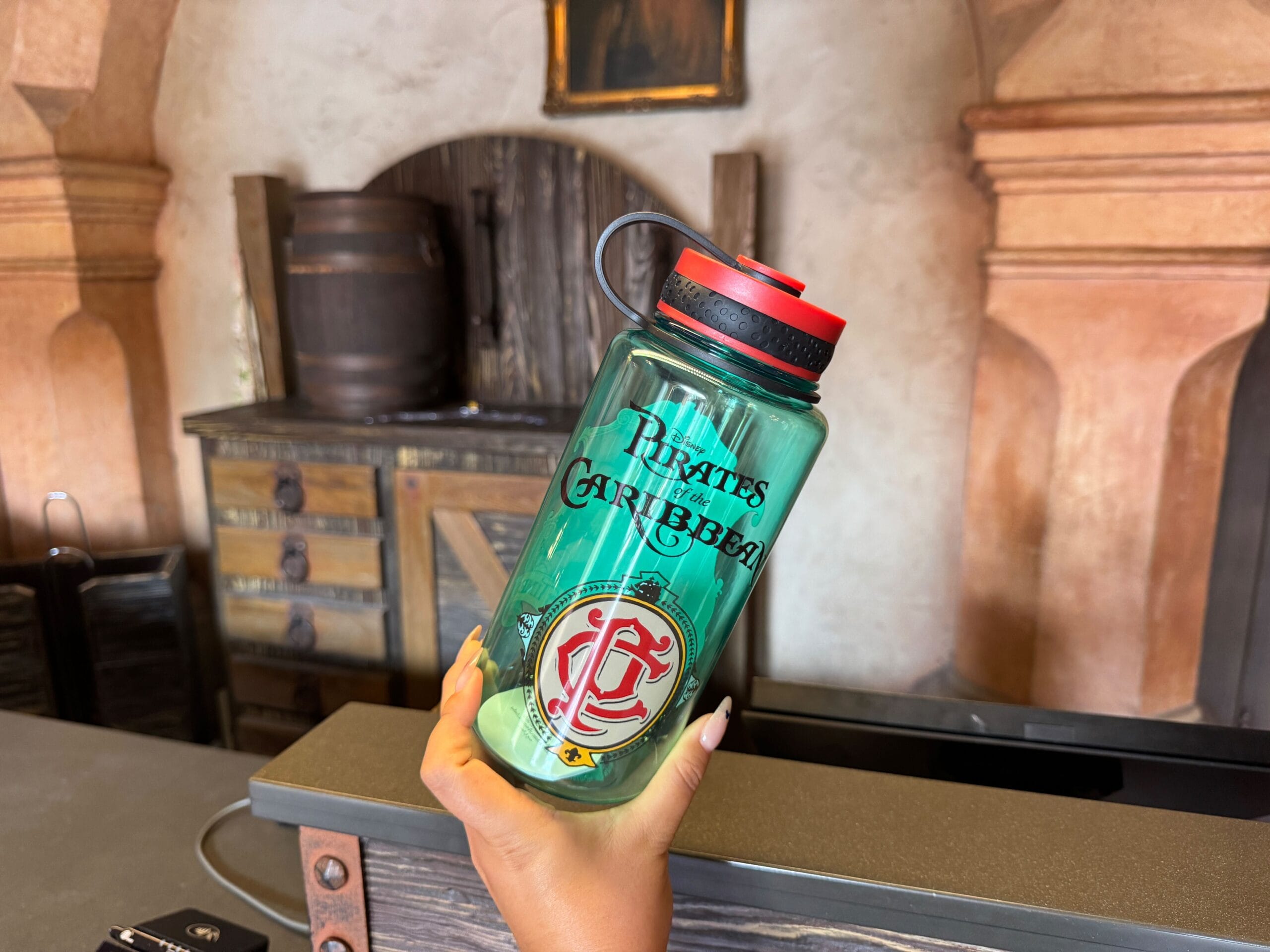 A hand holds a green "Pirates of the Caribbean" water bottle in a rustic, pirate-themed room.