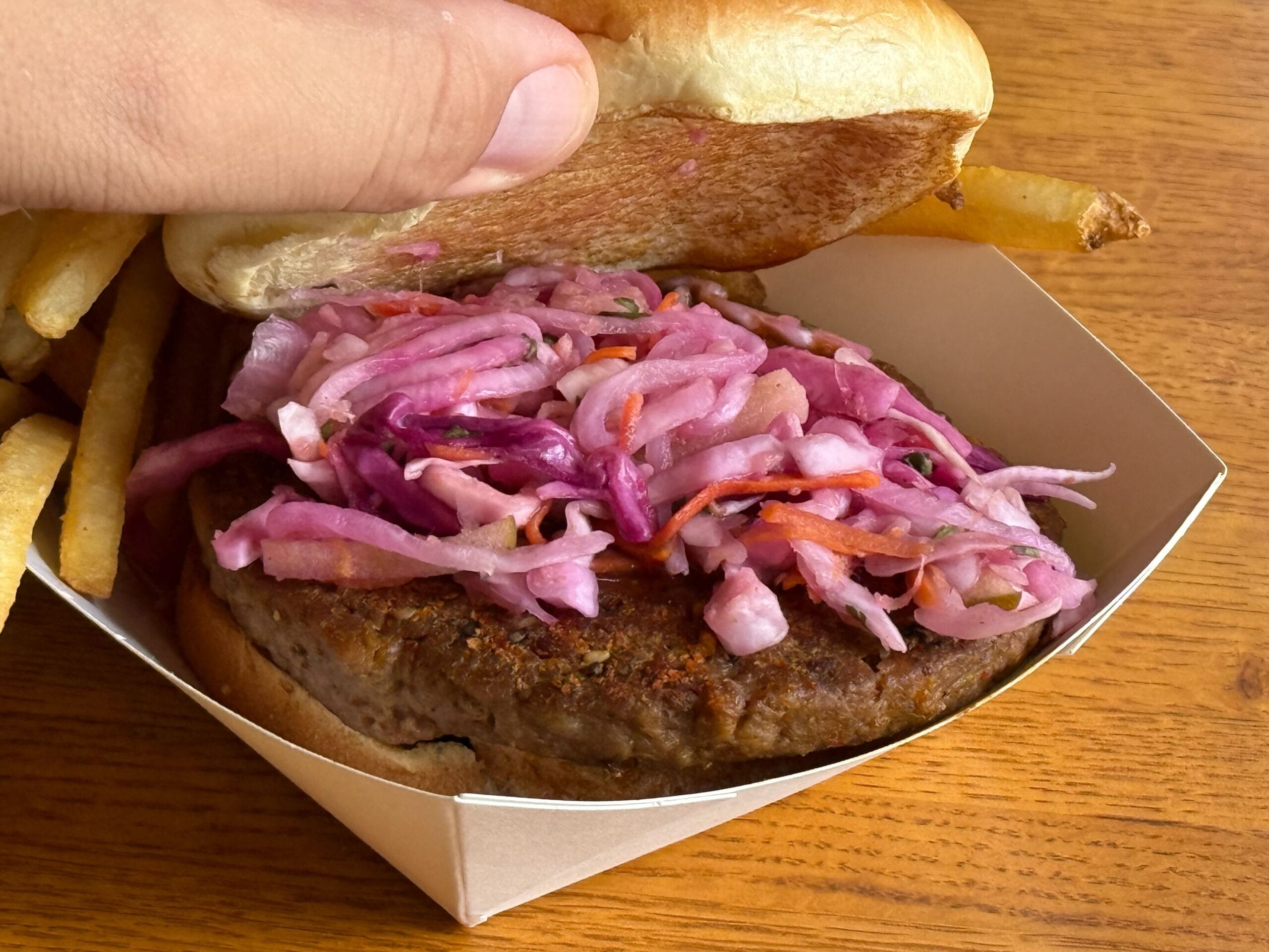A hand lifts the top bun of a burger with coleslaw and fries at Disney's Port Orleans Resort on a wooden table.
