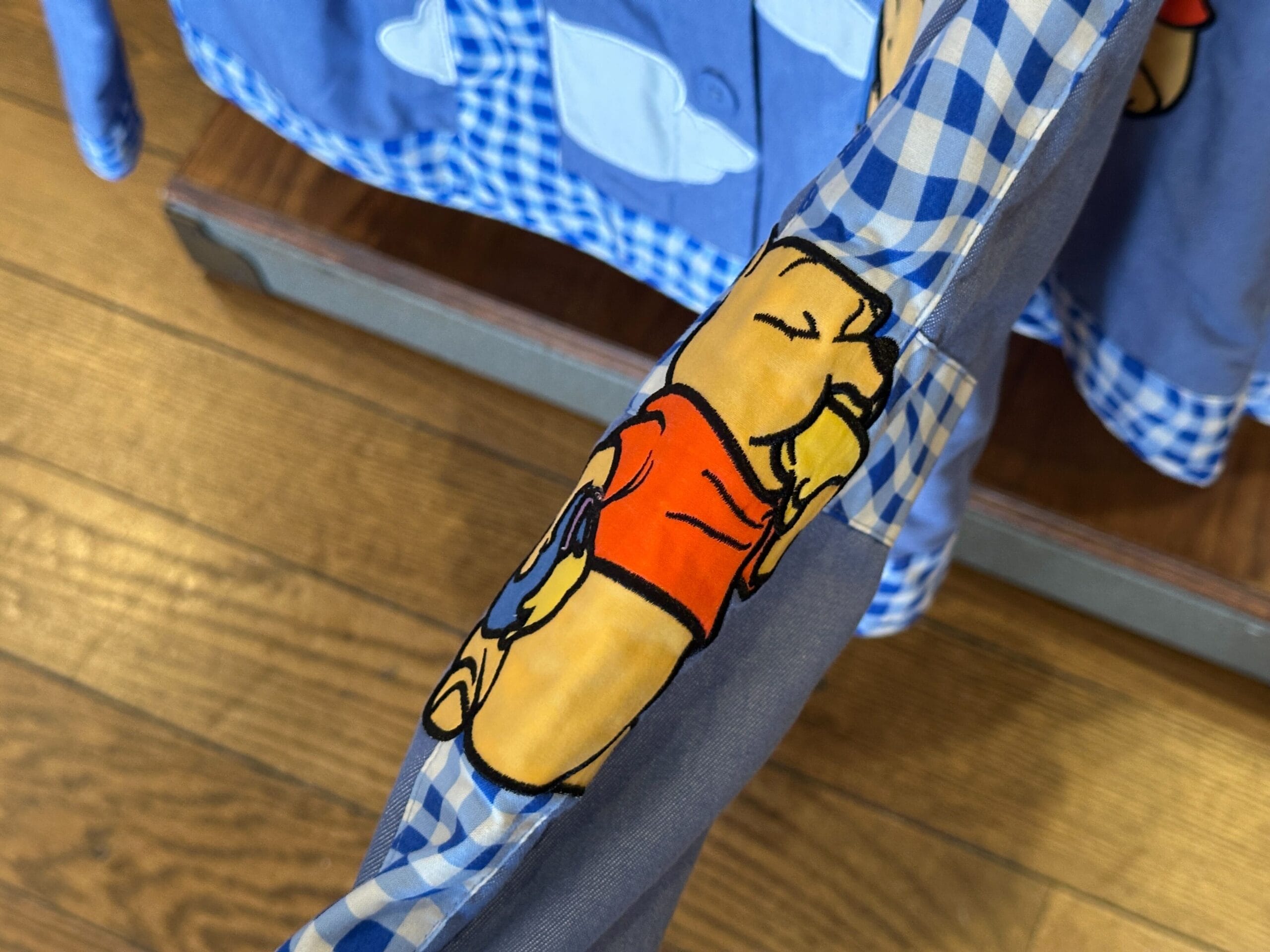 Close-up of a blue Winnie the Pooh sweater sleeve with cartoon graphic and checkered trim, on a wooden floor.