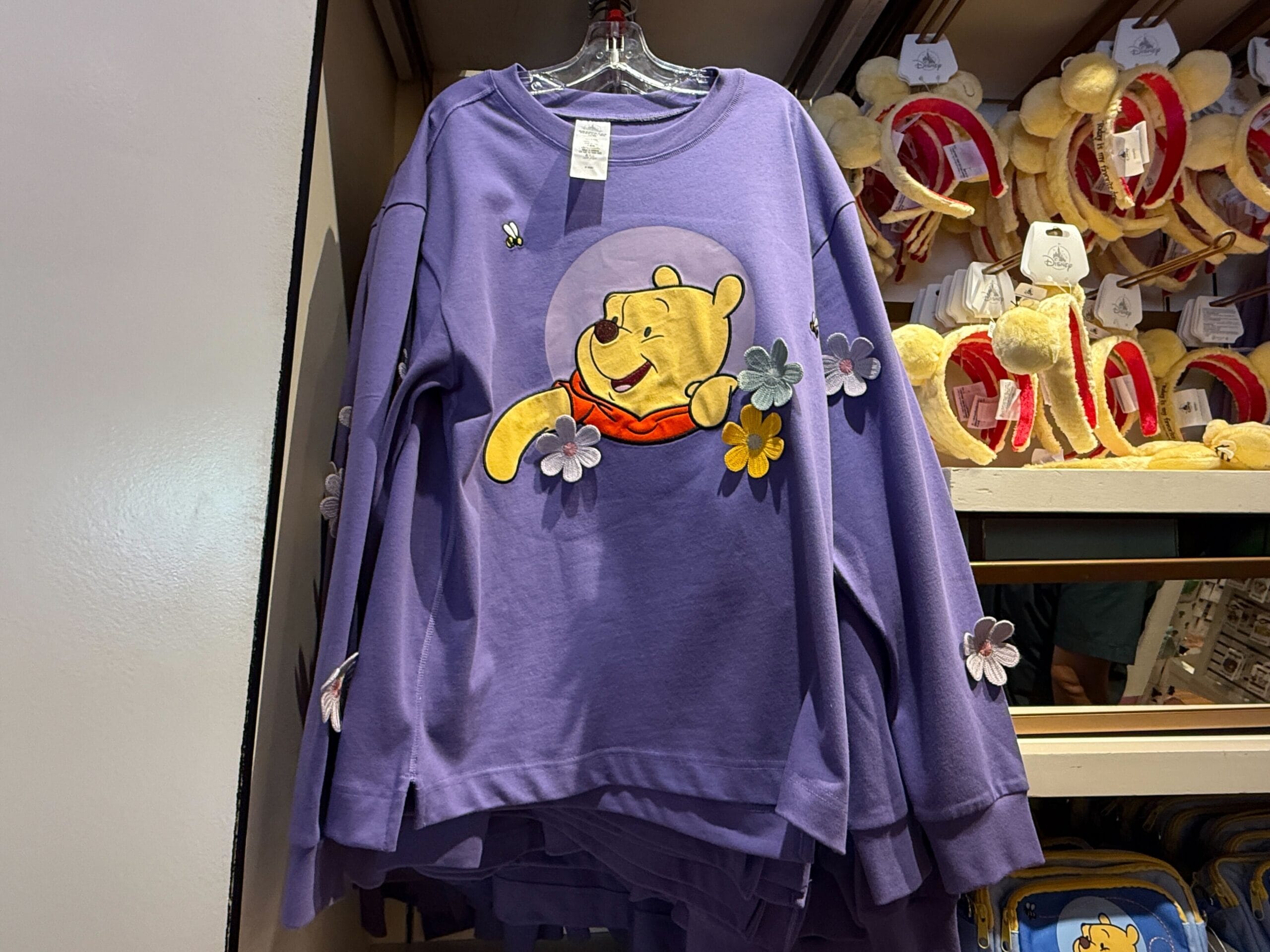 Purple winnie the pooh sweater with flowers and a bee, displayed on a rack beside Pooh ear headbands.