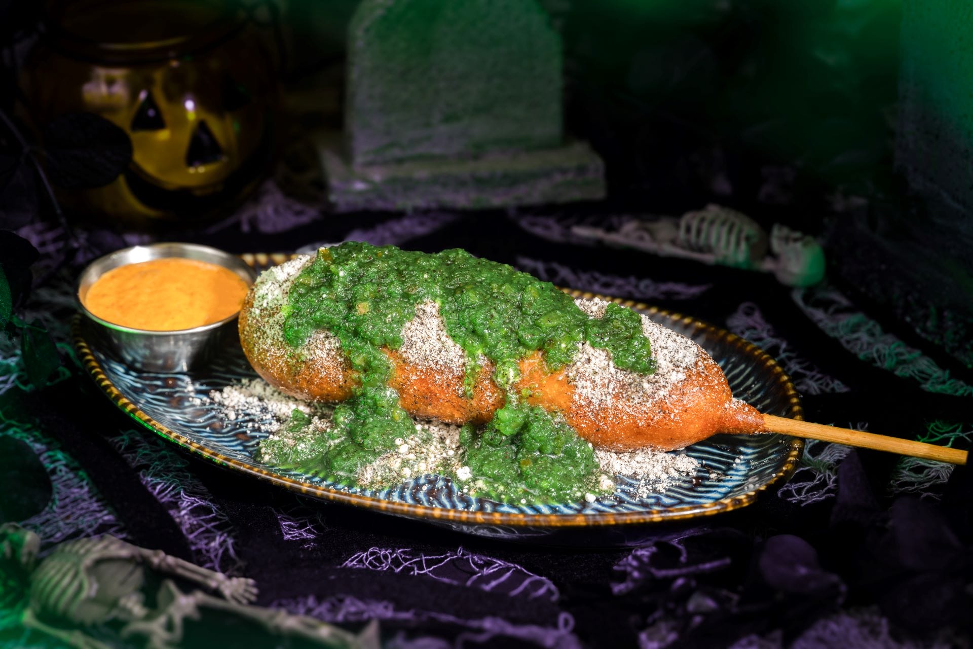 A festive corn dog with green sauce and cheese, served at Mickey's Not So Scary Halloween Party on a themed plate.