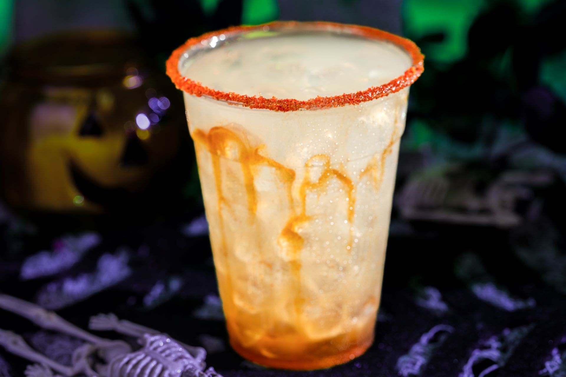 A spooky beverage with orange rim and syrup drips, served on ice, surrounded by Halloween decorations.