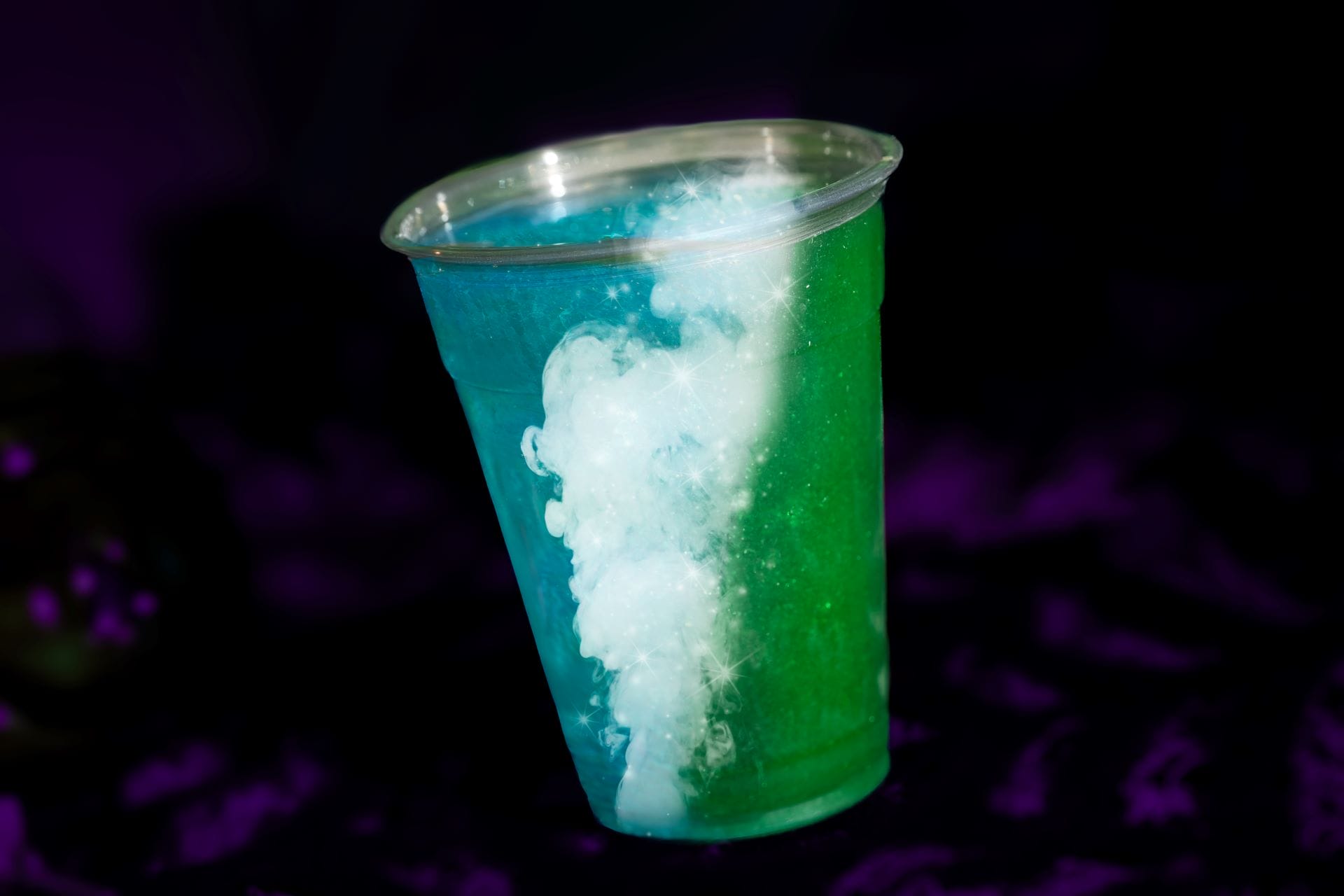A plastic cup of blue and green beverages with white swirling clouds, set against a dark purple background.