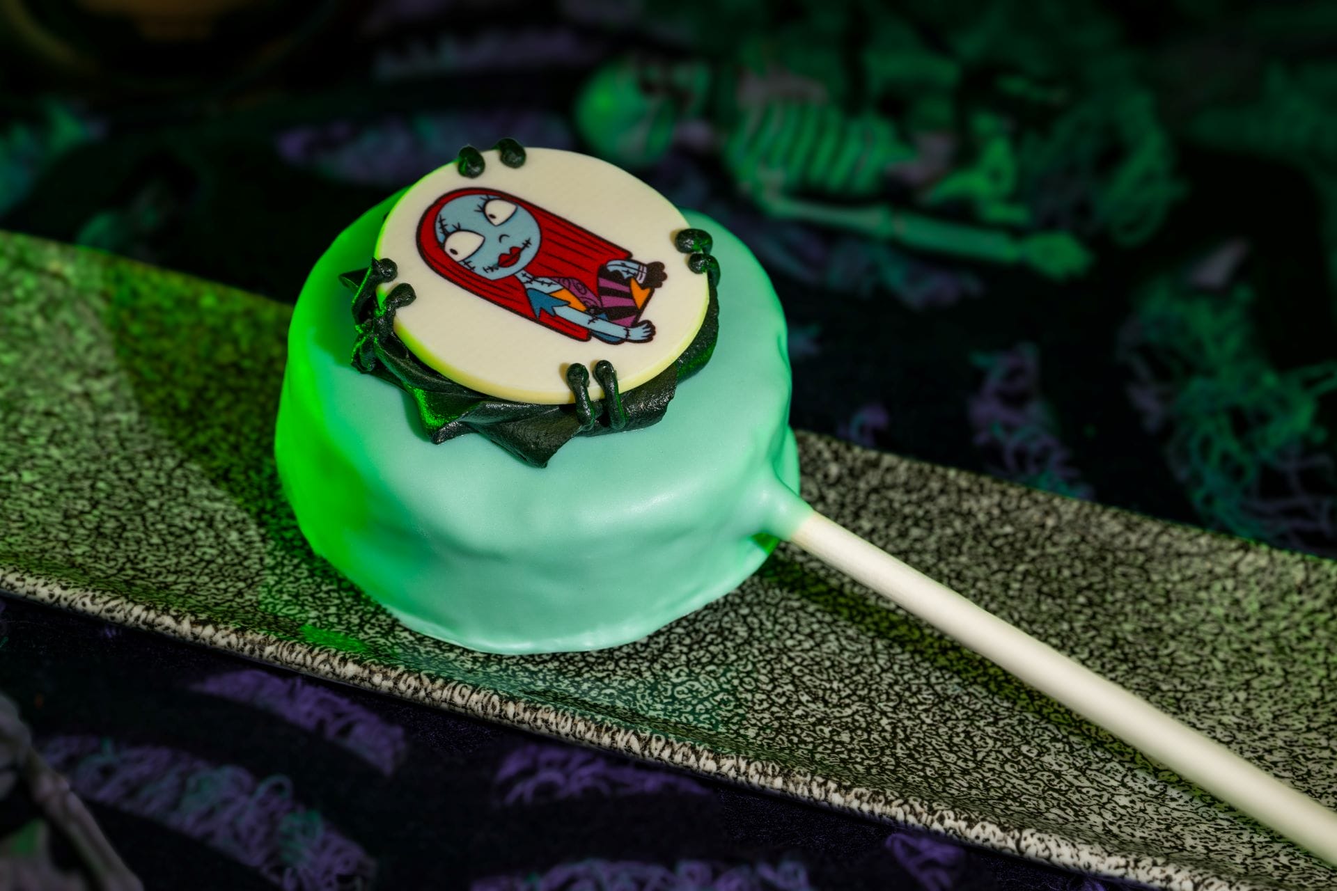 A round cake pop with pale green icing, inspired by Mickey's Not So Scary Halloween Party, sits on a textured tray.