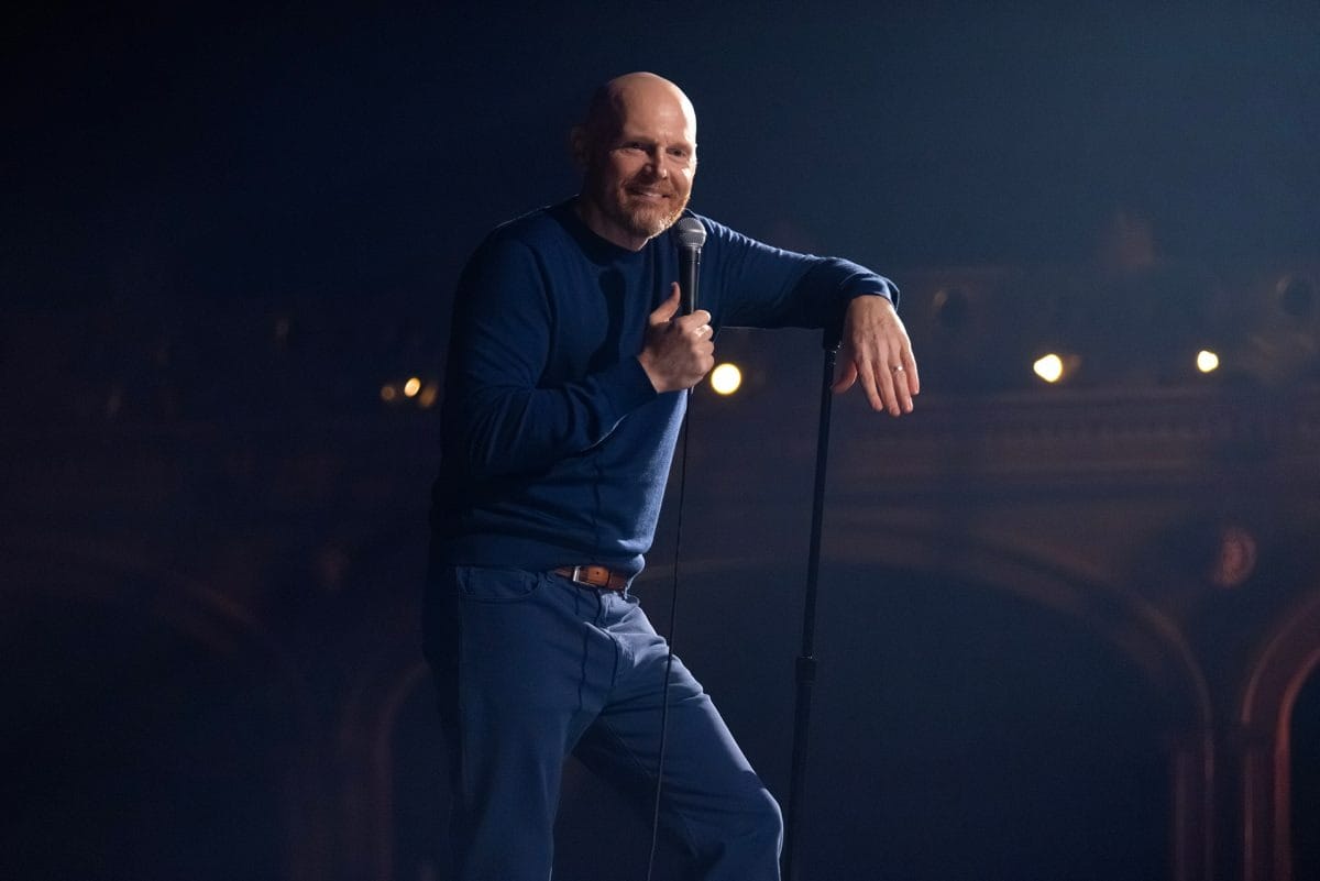 A man in blue holds a microphone and smiles while performing on stage at a Disney event under warm lighting.