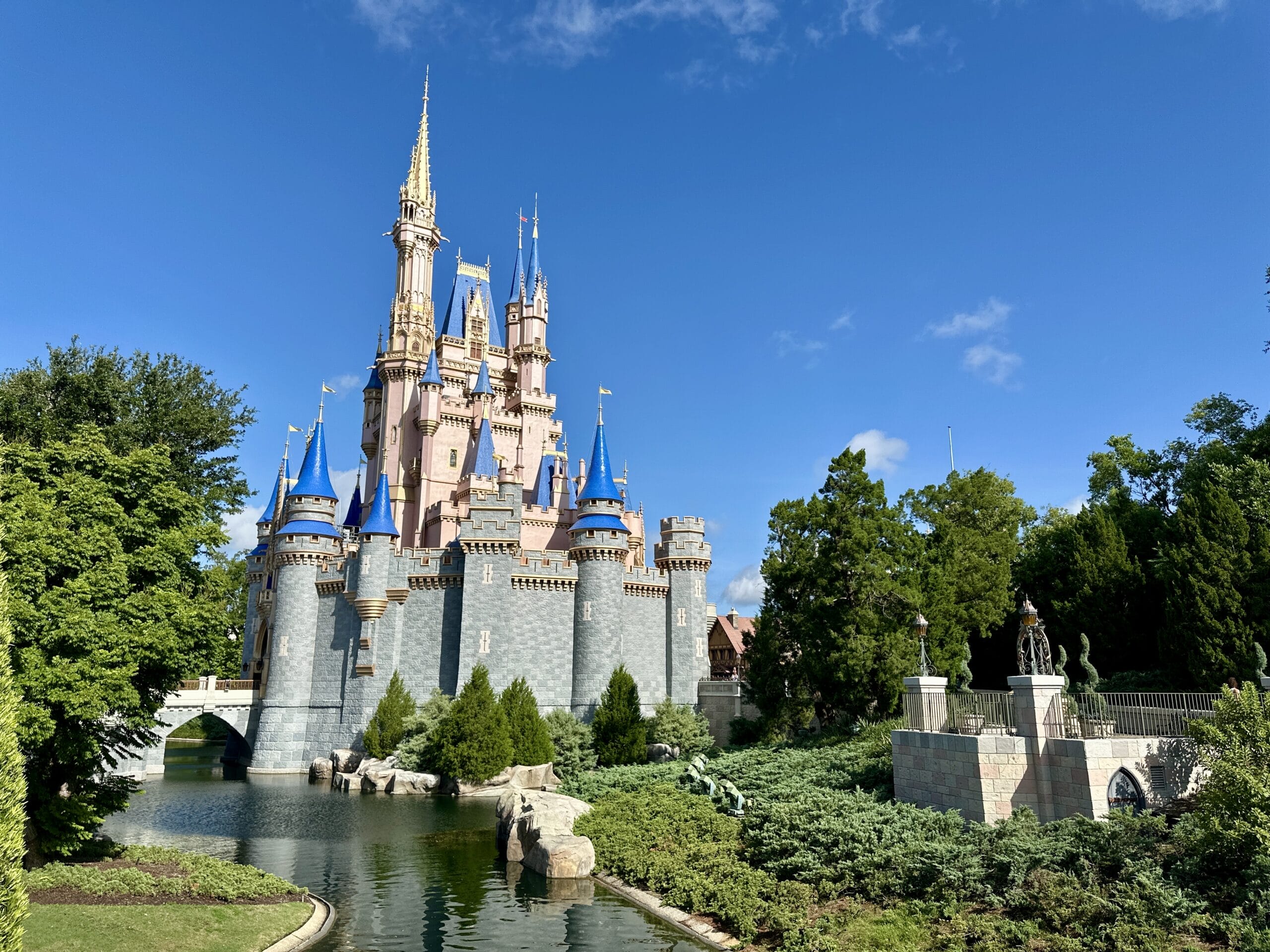 A fairytale castle with blue and gold spires stands in Magic Kingdom, surrounded by greenery and a sparkling moat.