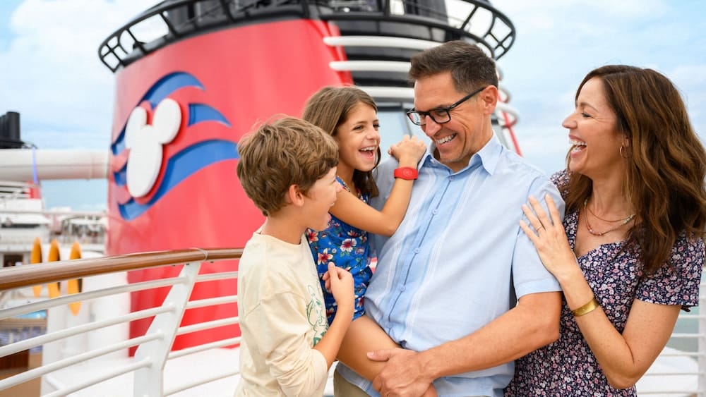 Smiling family stands on a cruise ship deck with a large red funnel featuring a Mickey Mouse logo behind them.
