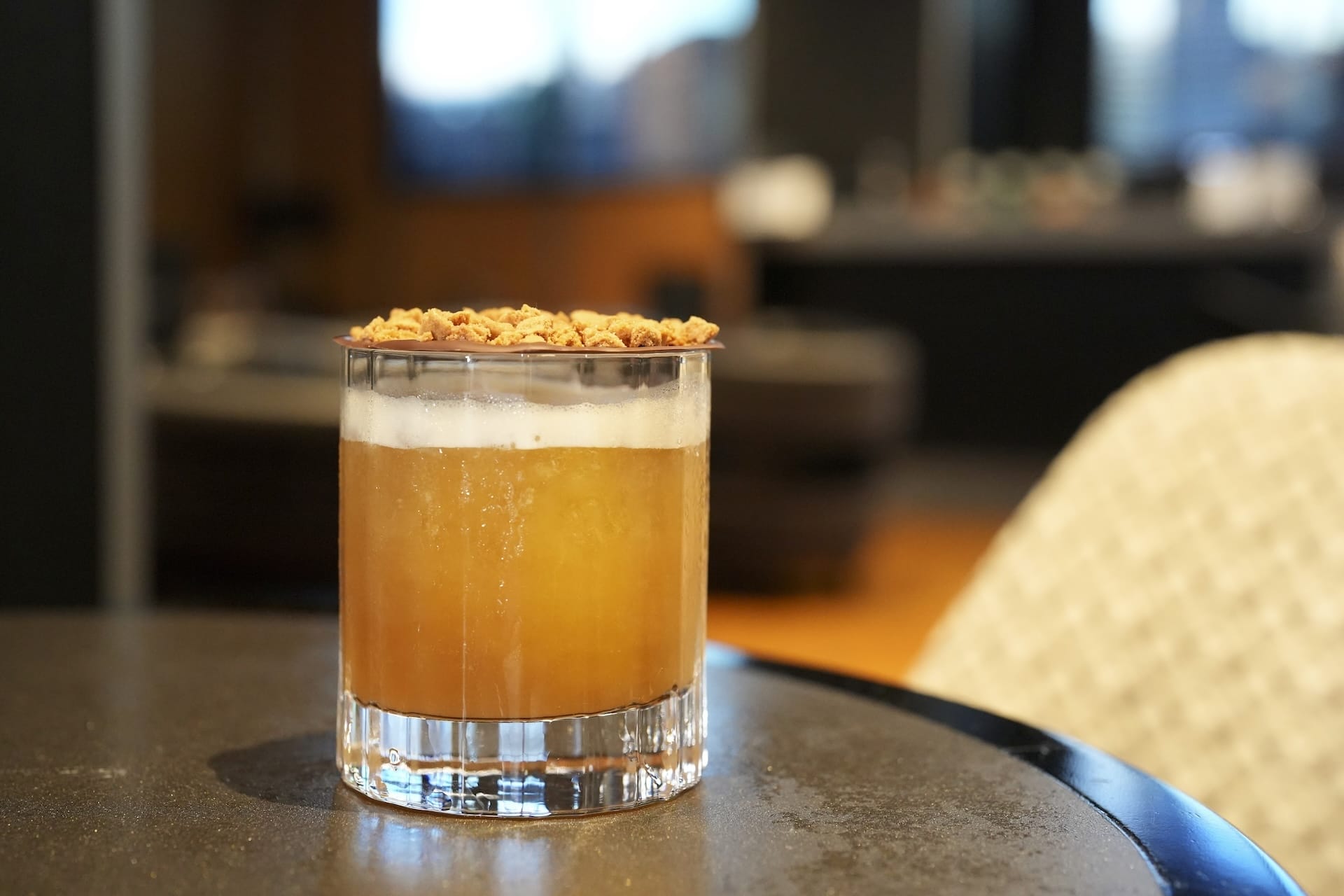 A glass of orange-brown cocktail with foam and granola sits on a dark table, inspired by Fantastic Four food and beverages.