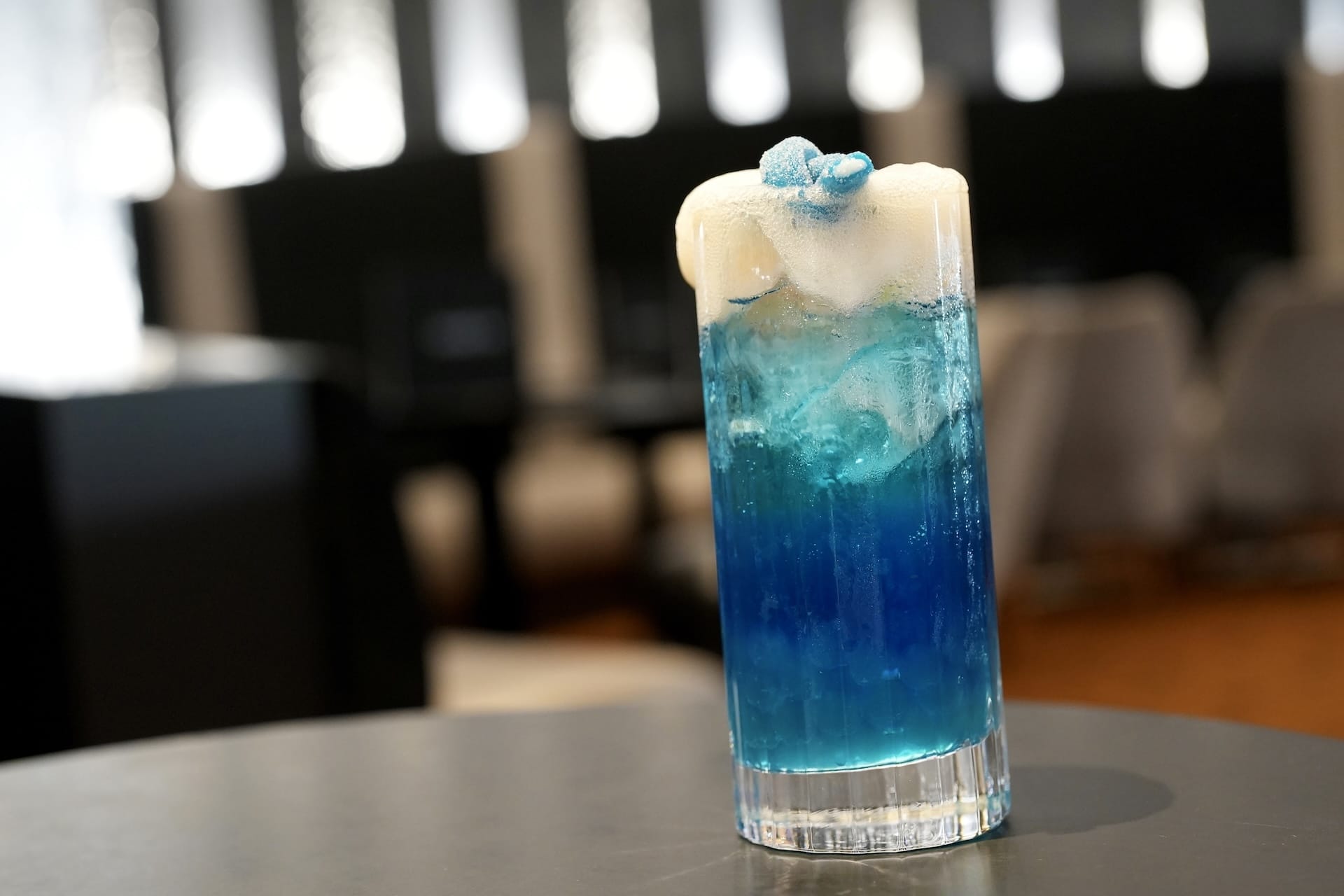 A tall glass of blue cocktail with ice and foam, garnished with a blue candy, channels a Fantastic Four vibe on a dark table.