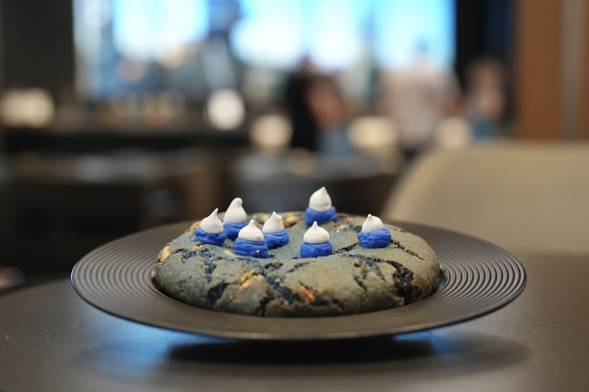 A large blue cookie inspired by the Fantastic Four sits on a black plate amid a blurred indoor setting.