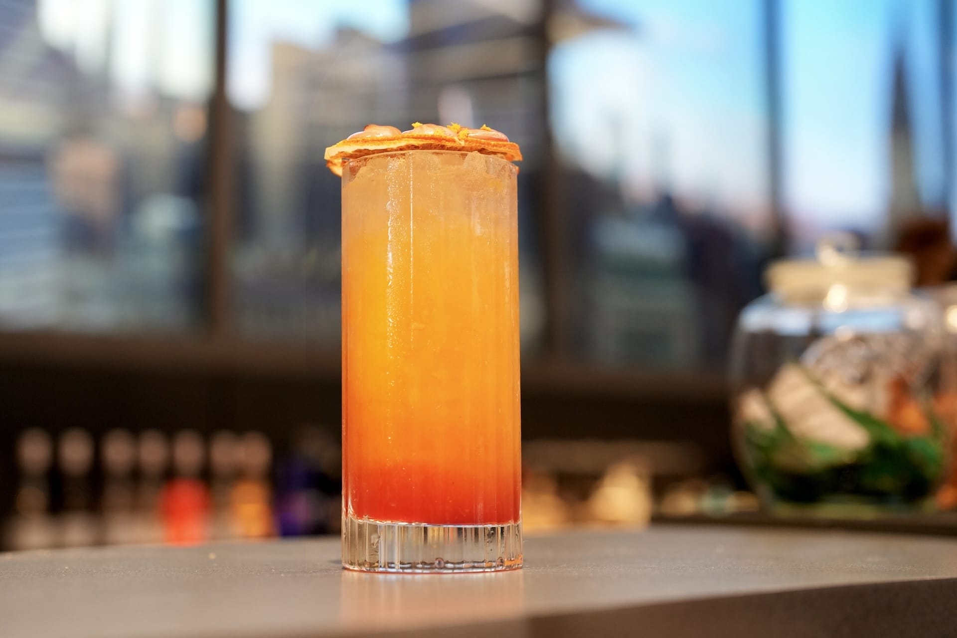 A tall glass of orange and red layered cocktail, inspired by Disney Parks food and beverages, sits on a countertop indoors.