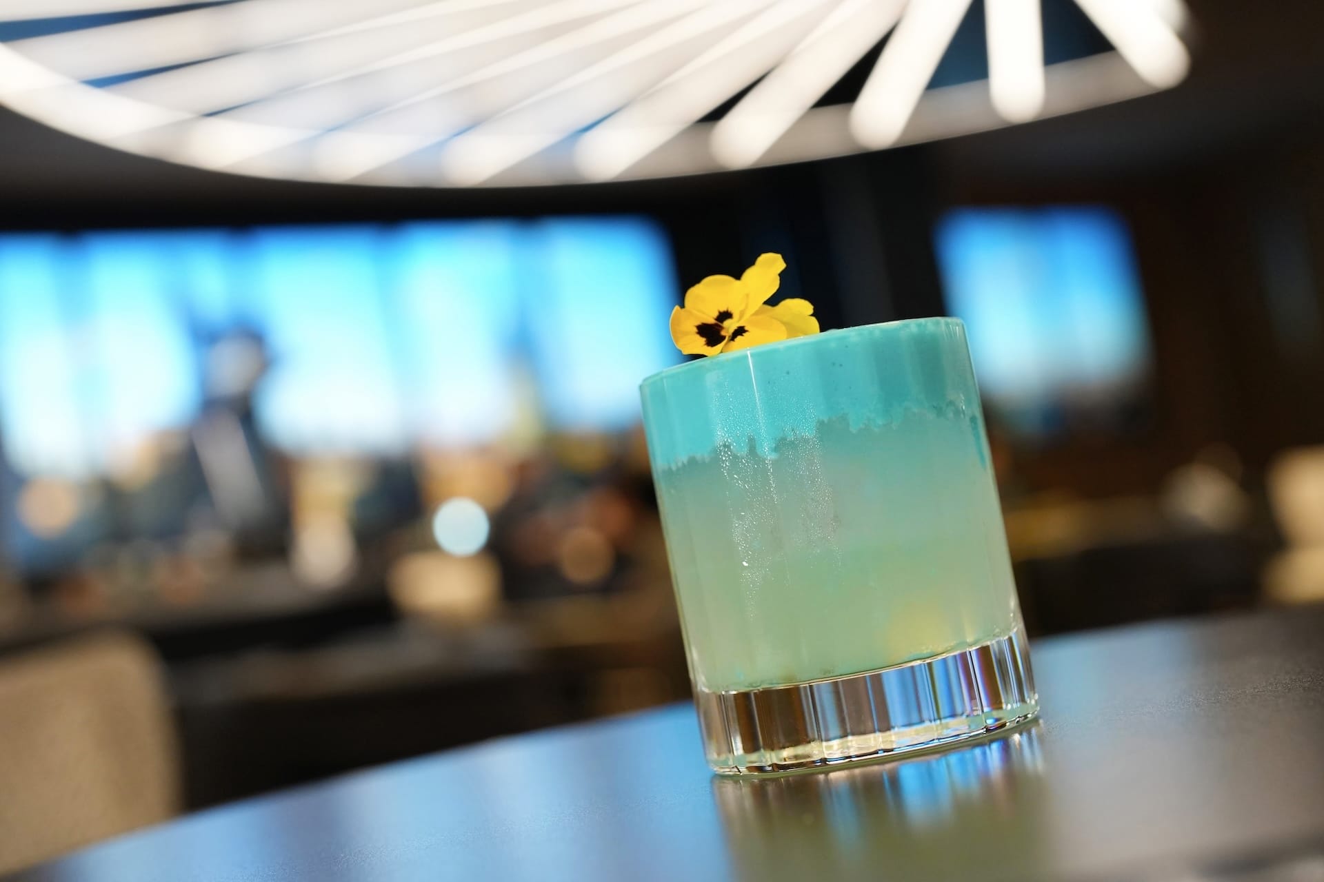 A blue and yellow Food and Beverages cocktail in a glass, garnished with a yellow flower, glows amid blurred lights.