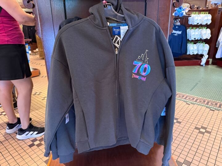 Gray Disneyland 70th anniversary zip-up hoodie celebrates the happy collection, hanging on a store rack with people and merchandise.