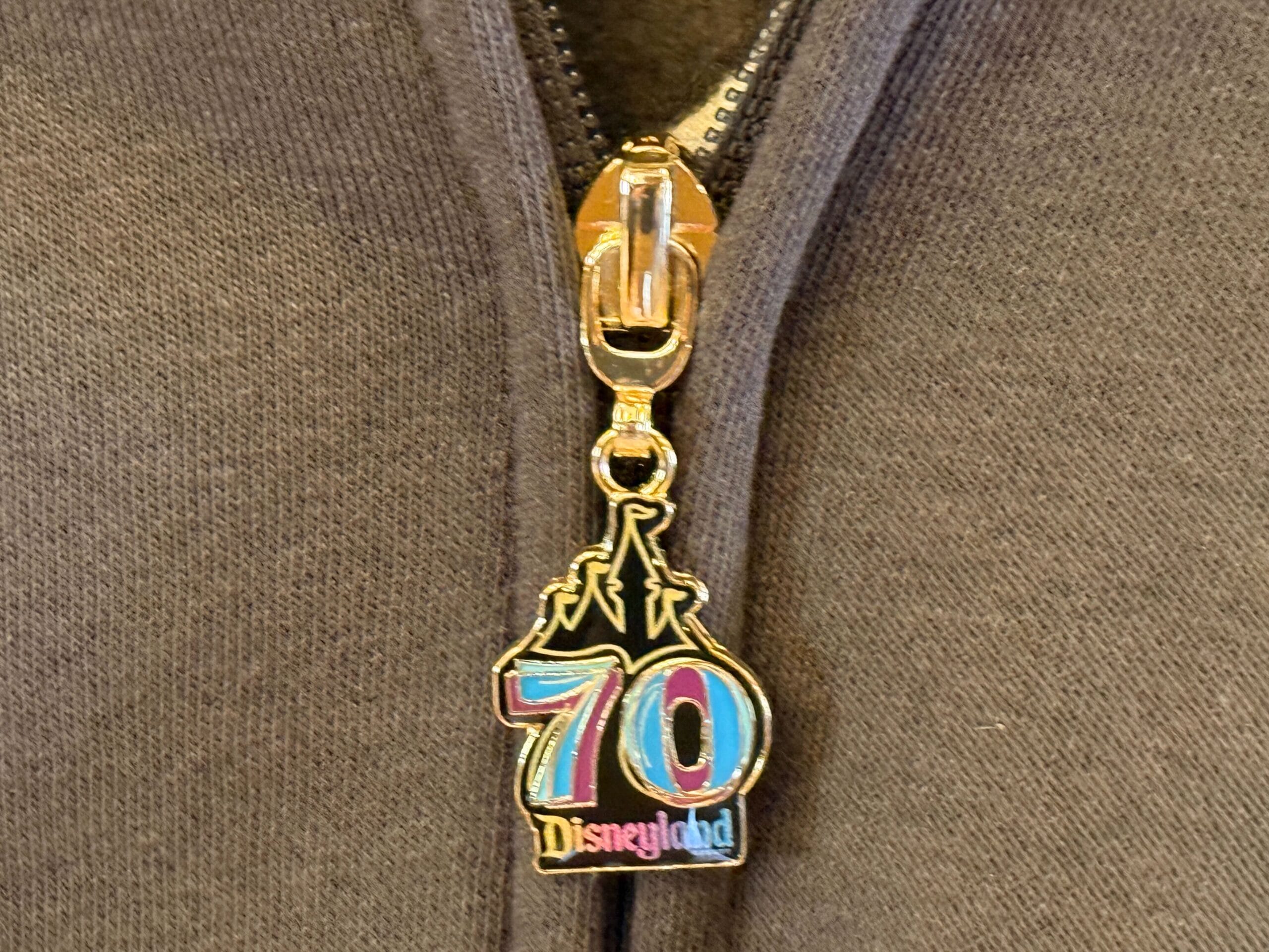 A zipper pull celebrates "70 Disneyland" in colorful text and castle, adding joy to any happy collection on a gray garment.