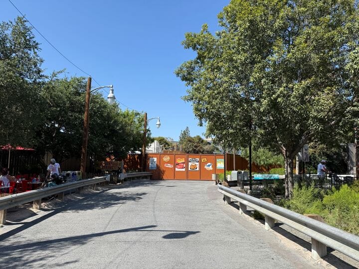 A paved path in DCA leads to construction walls with cartoon signs near Avengers Campus, surrounded by trees and seating.