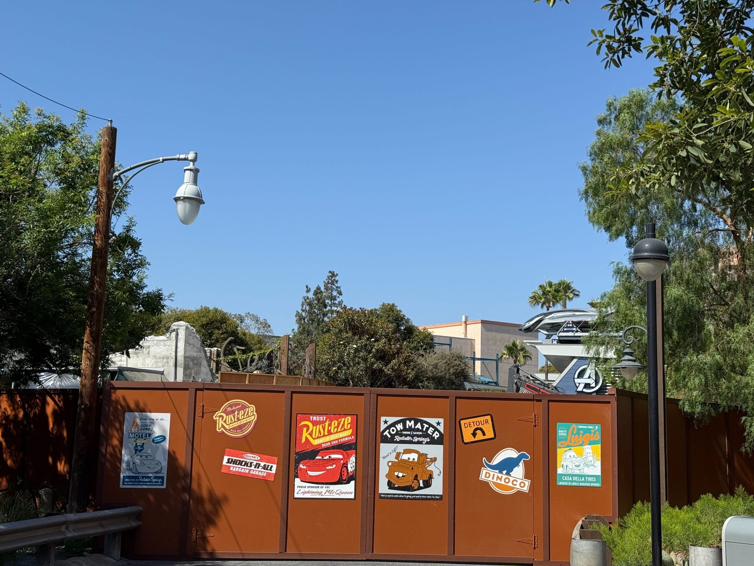 Brown construction fence with Cars-themed signs at DCA, trees, and updated Avengers Campus building in the background.