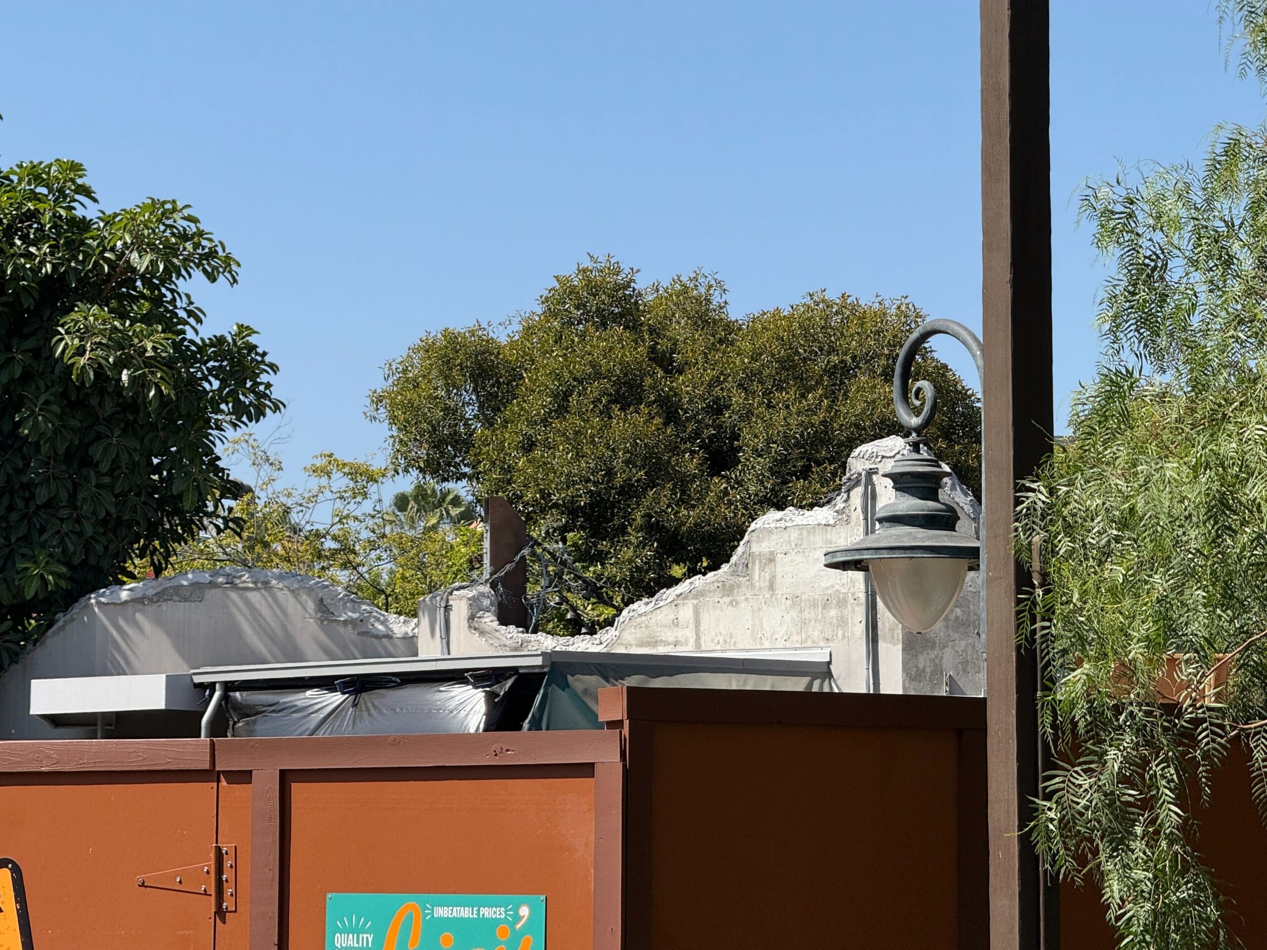 A partially demolished building at DCA behind a brown construction fence—an update near Avengers Campus, with trees and a streetlamp.