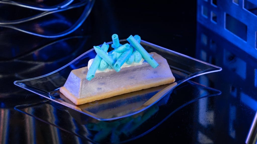 A rectangular dessert with blue garnish, inspired by Disney Parks, sits on a clear plate atop a glossy black surface.