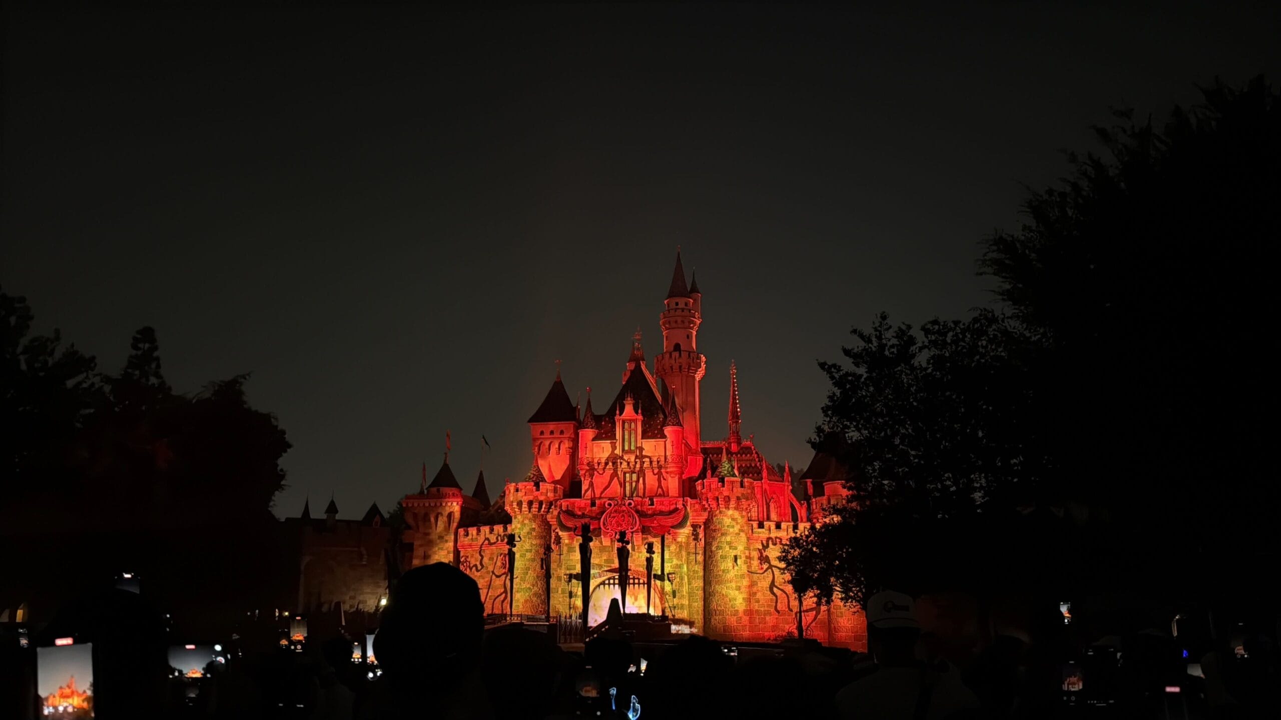 Disneyland castle illuminated in red and orange lights at night, celebrating America fireworks with silhouettes in the foreground.