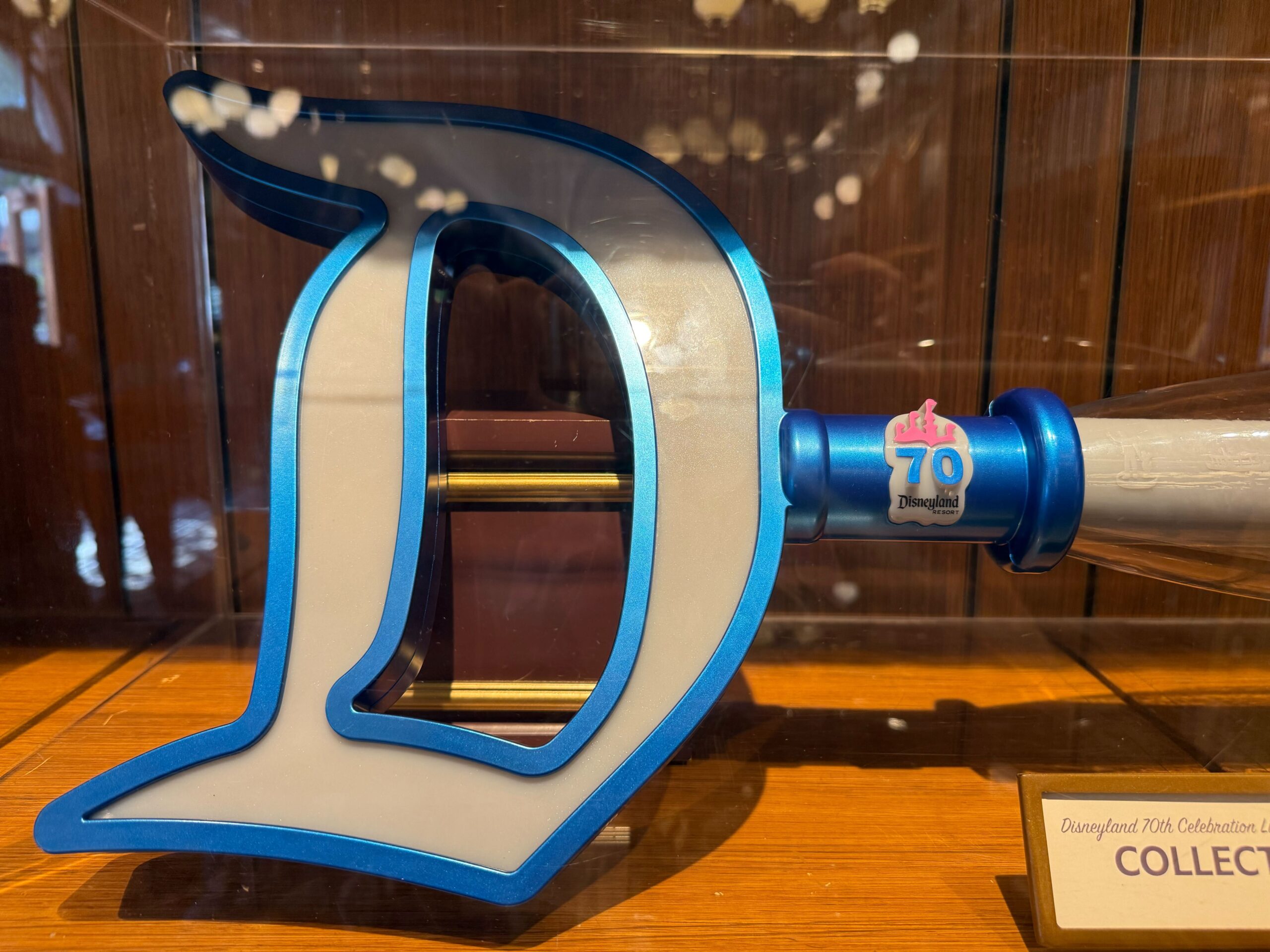 A blue and white "D" Disneyland 70th Collectible Key displayed with a Celebration logo on a wooden shelf.