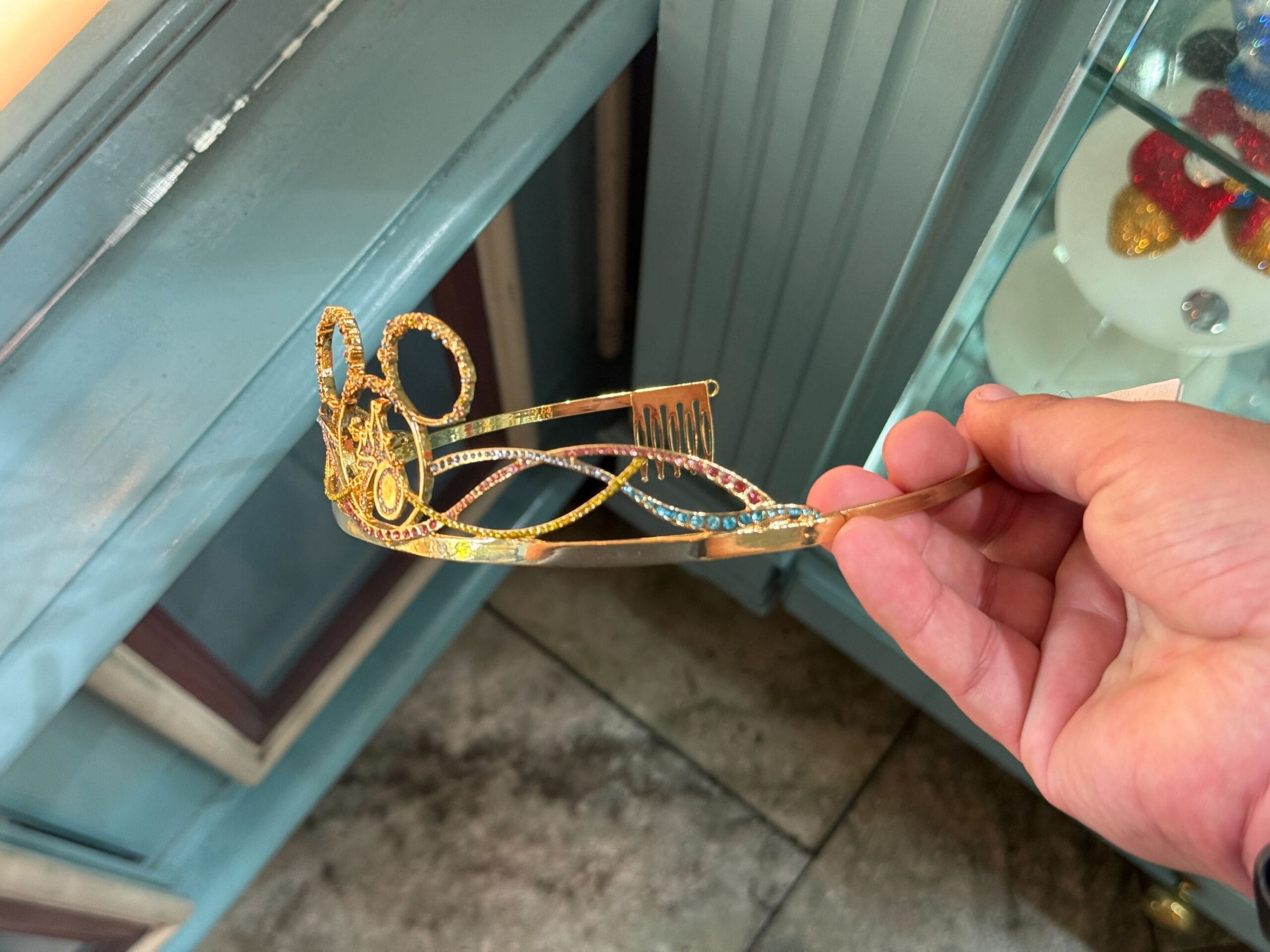 A hand holds a gold, jeweled tiara with intricate designs and blue and pink gems inside a shop.