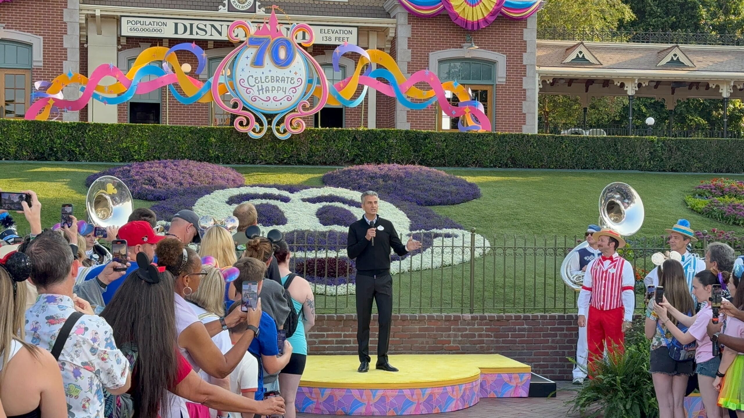Bob Iger speaks on stage at Disneyland 70th Anniversary, surrounded by cheerful crowds and festive decorations.