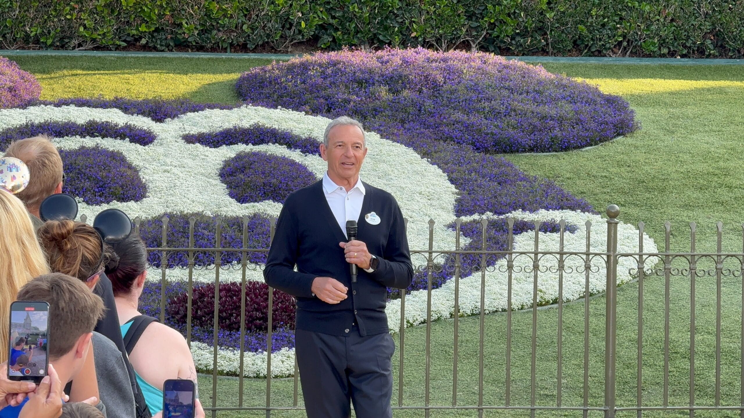 Bob Iger with a microphone speaks to a small crowd at Disneyland’s Mickey Mouse-shaped flower bed.