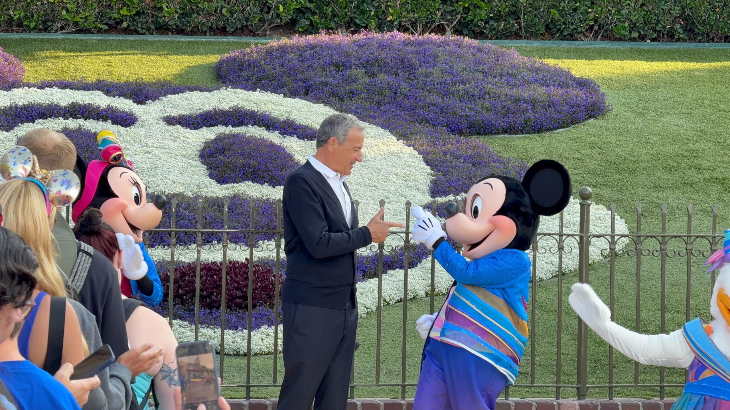 Bob Iger and Mickey Mouse gesture to each other by a floral Mickey at Disneyland's 70th Anniversary, with visitors nearby.
