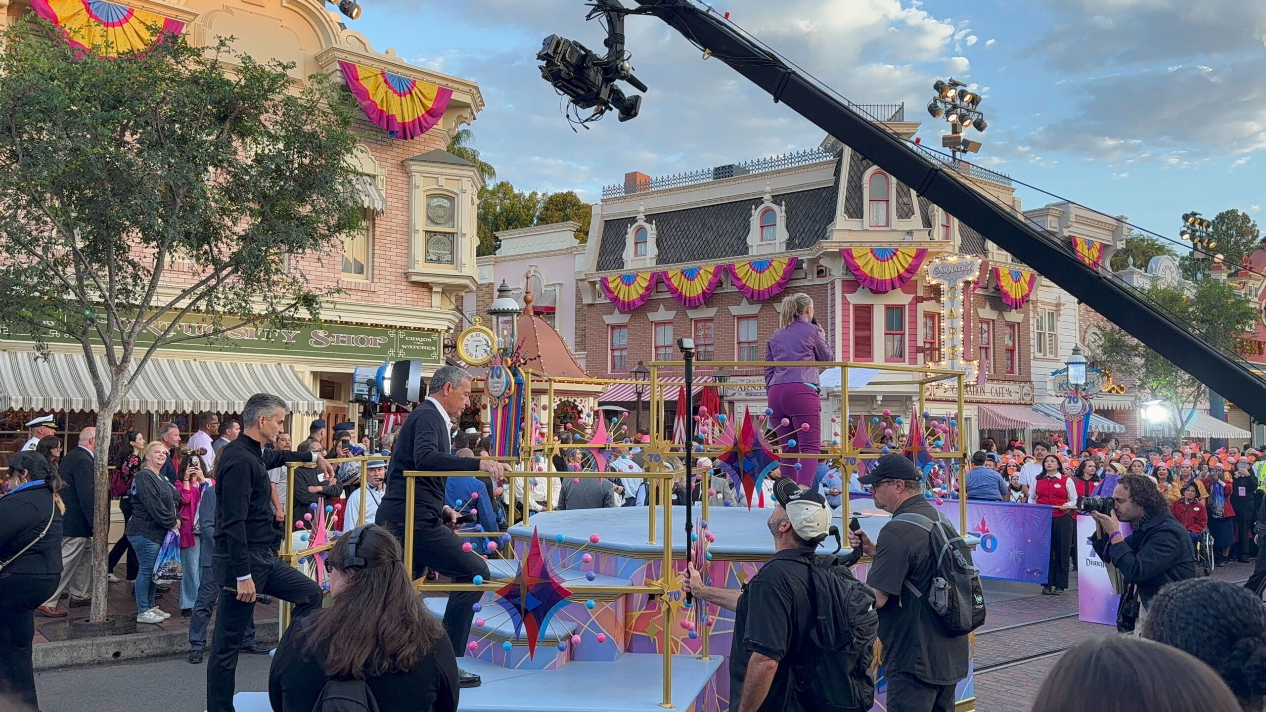 A parade float with performers on Main Street at Disneyland’s 70th Anniversary; camera crew films amid festive crowds and banners.
