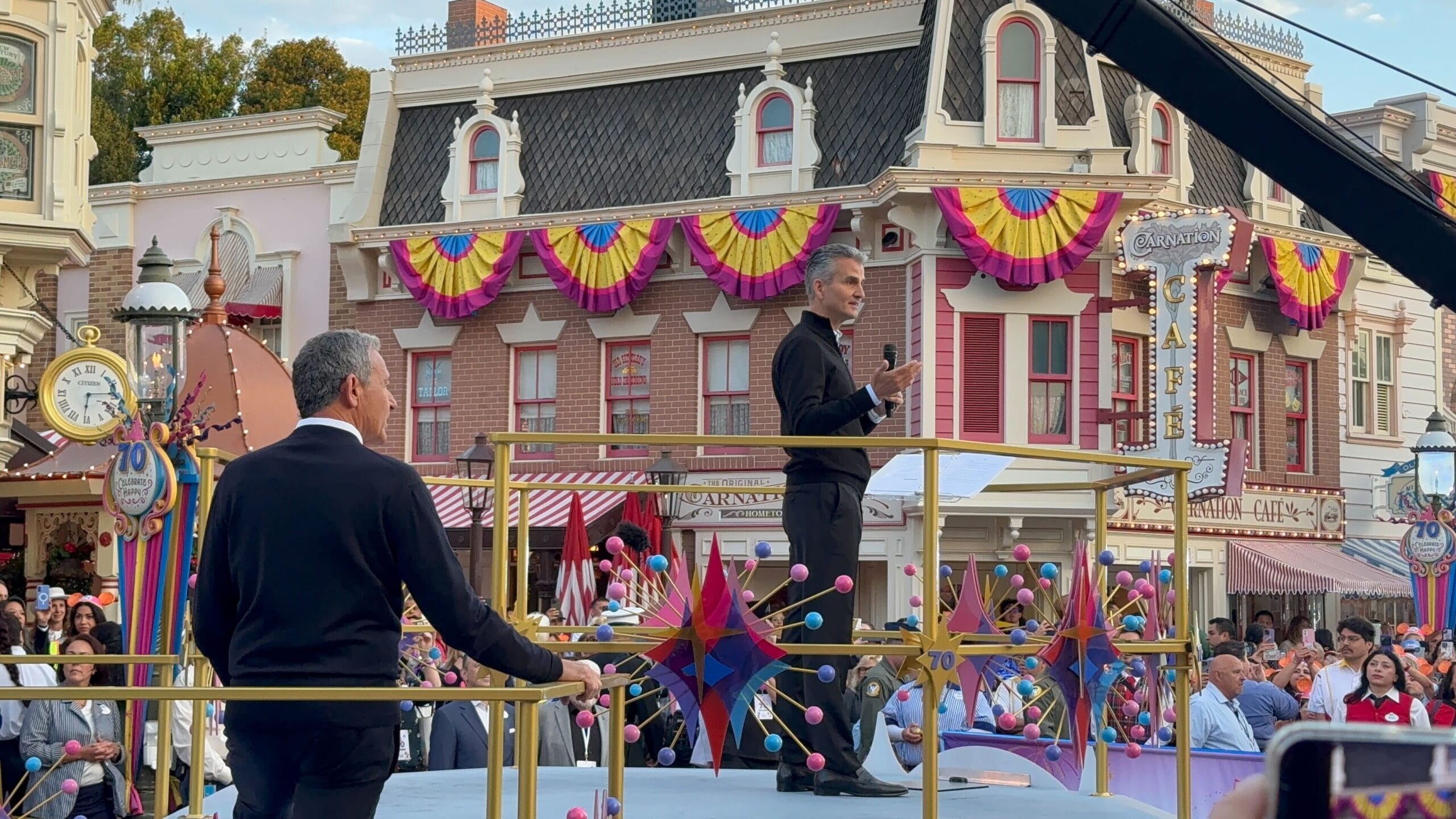 Two men stand on a decorated parade float during Disneyland's 70th Anniversary, in front of Main Street’s Victorian-style building.