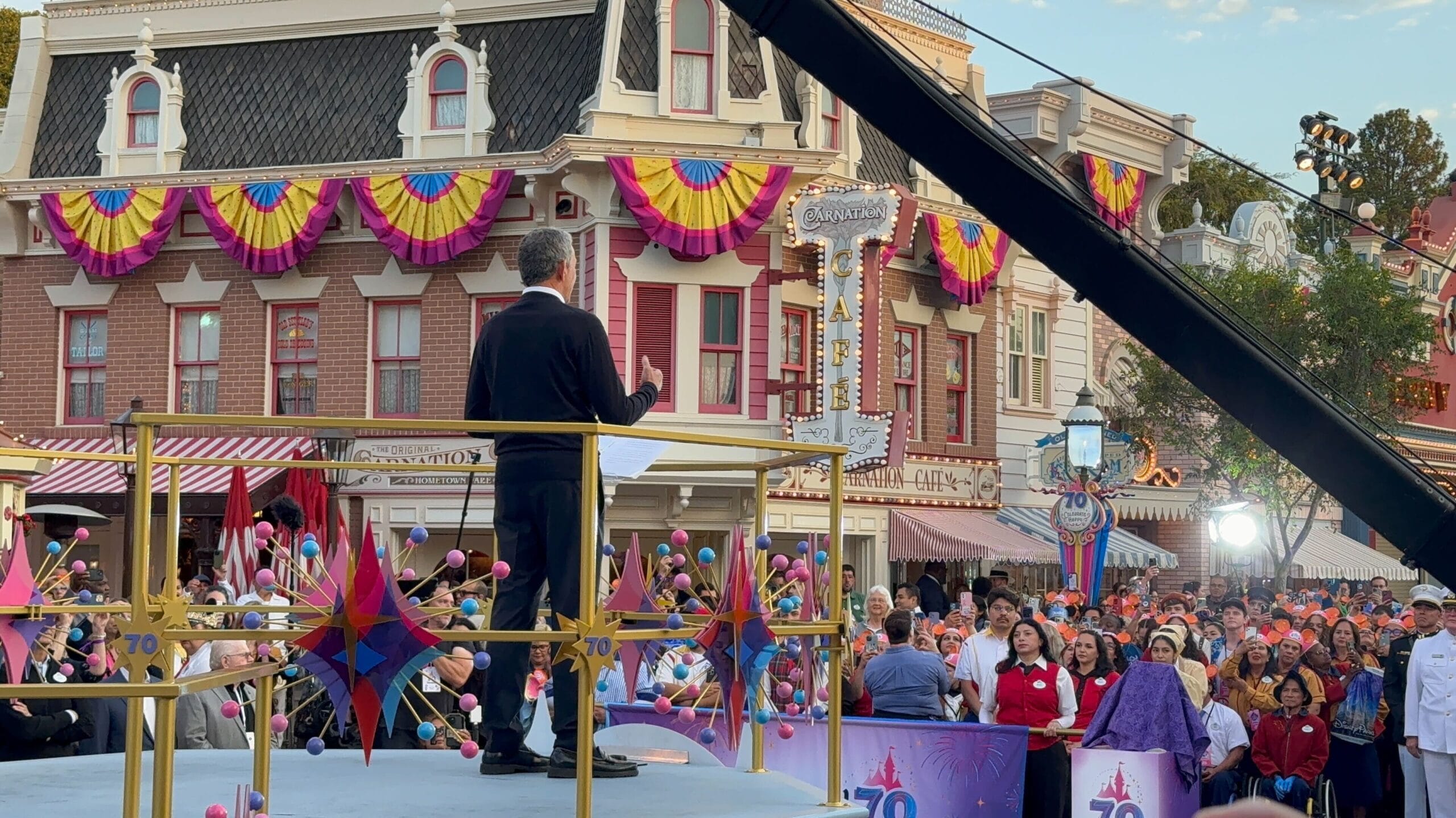 A person speaks on stage to a large crowd at a festive outdoor ceremony during Disneyland's 70th Anniversary.