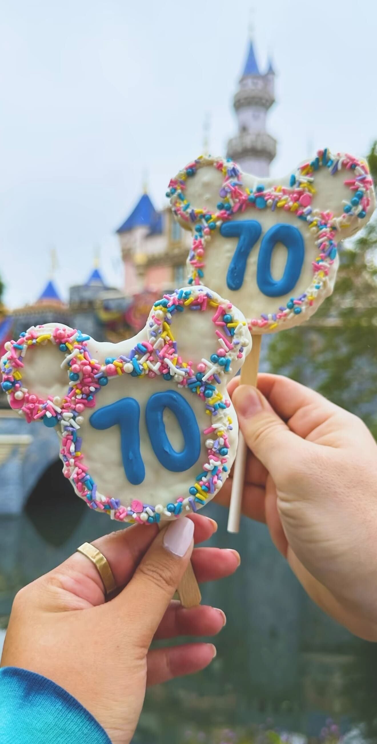 Two hands hold Mickey-shaped new treats with "70" in blue icing and sprinkles, celebrating Disneyland 70th Anniversary.