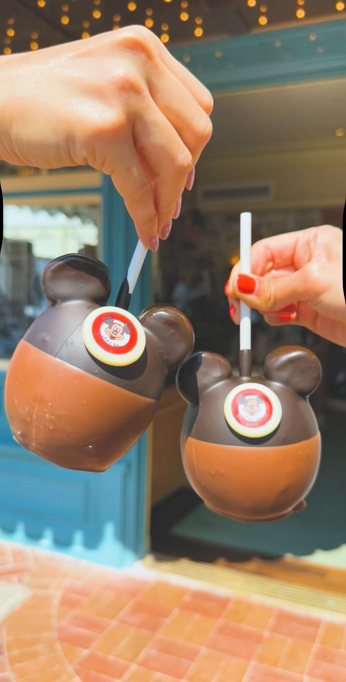 Two hands holding new treats: Mickey-shaped chocolate cups with straws, decorated with Captain America shields.