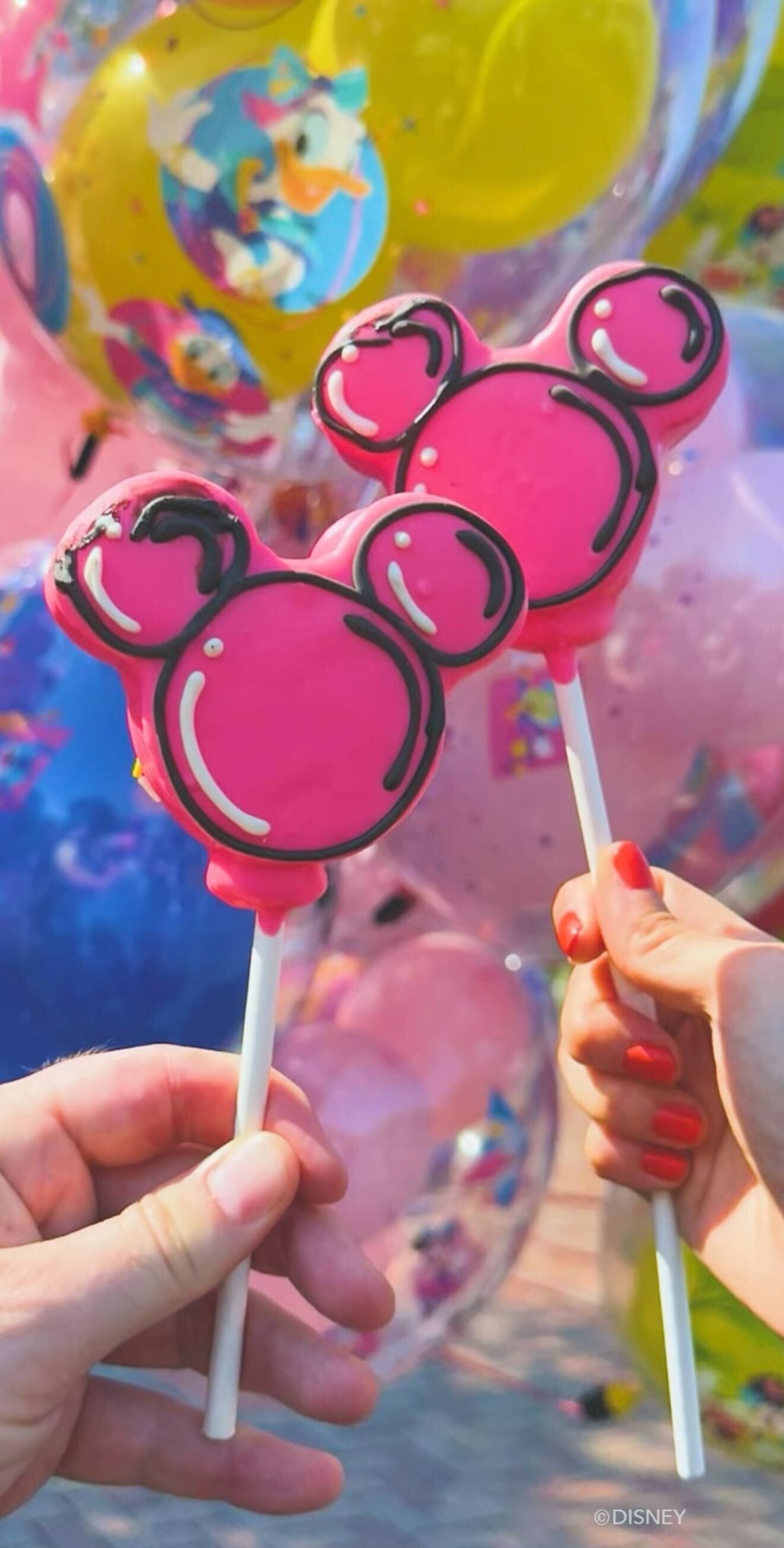 Two hands hold new DOLE Whip treats on sticks, with colorful Disney balloons at Disneyland 70th Anniversary in the background.