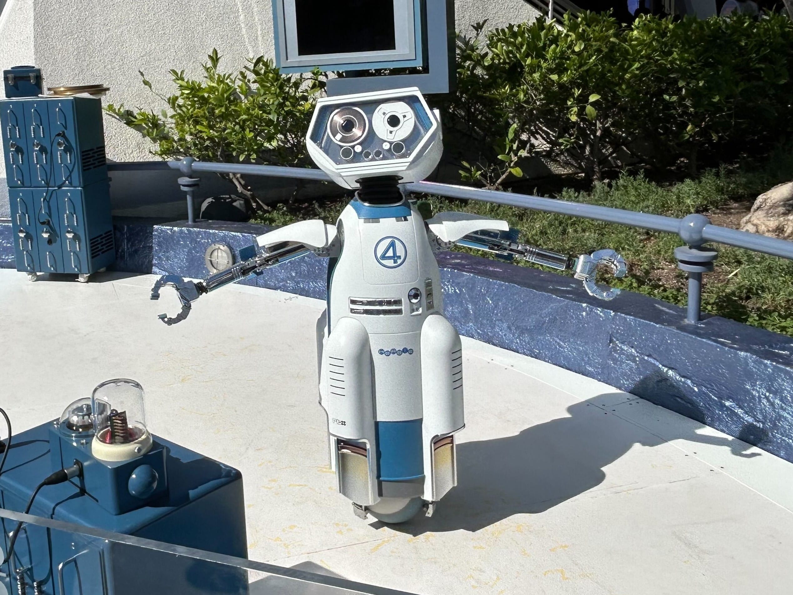 A white and blue HERBIE robot with claw arms stands outdoors by a curved blue wall at Disneyland.