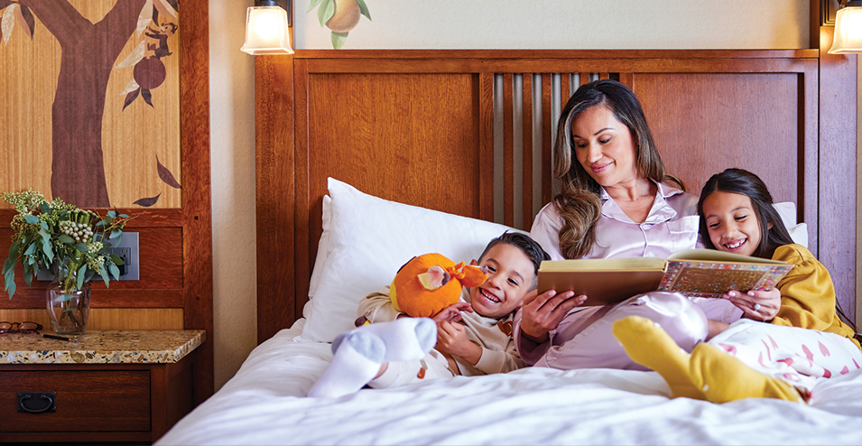 A mother reads a book to her smiling son and daughter while sitting together on a cozy bed.