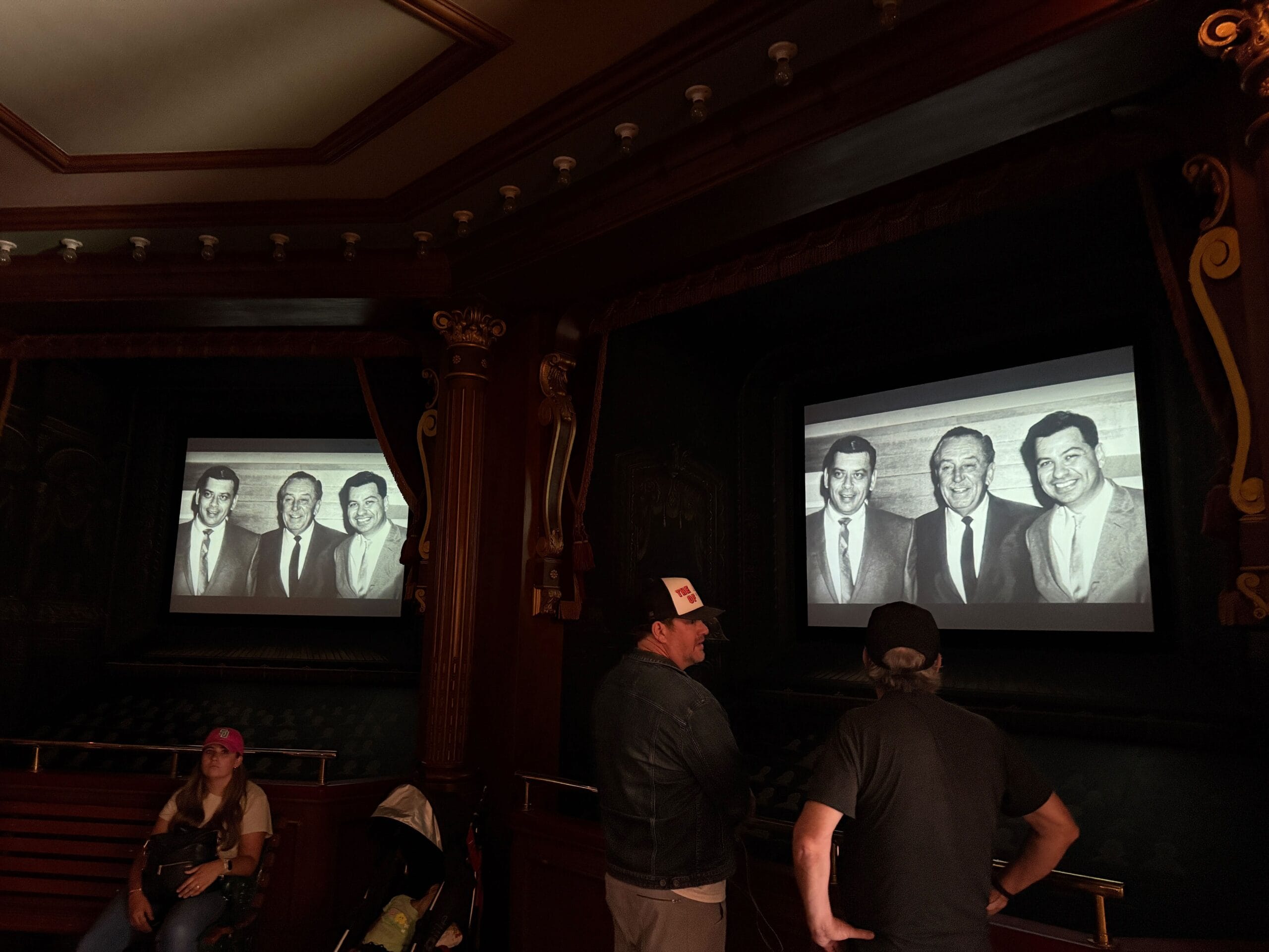 Two men face a screen at Main Street Cinema showing a photo; a woman sits nearby at the Sherman Brothers Tribute.