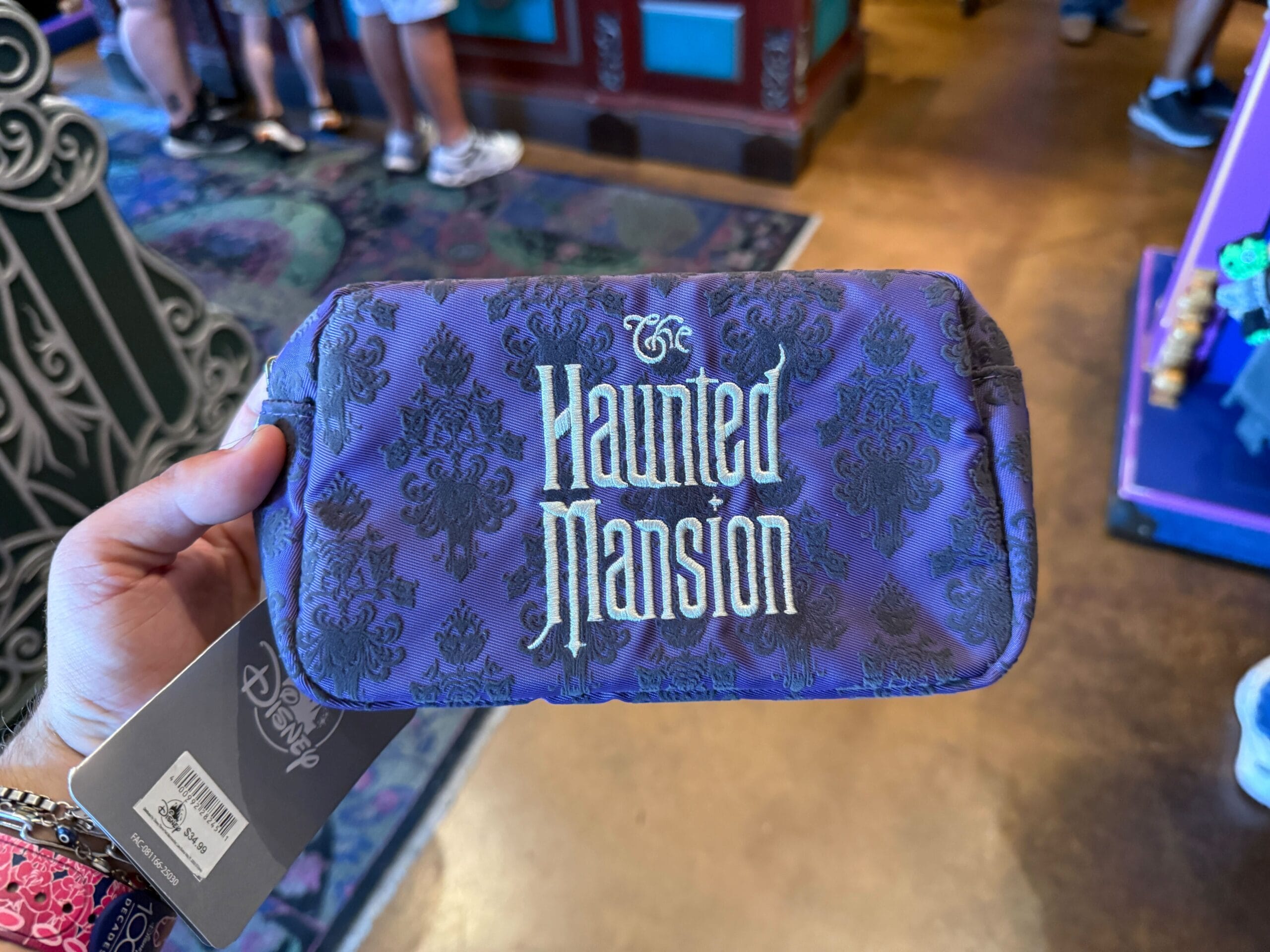 A hand holds a purple pouch with "The Haunted Mansion" text, classic Disneyland HM Merchandise in a themed store.