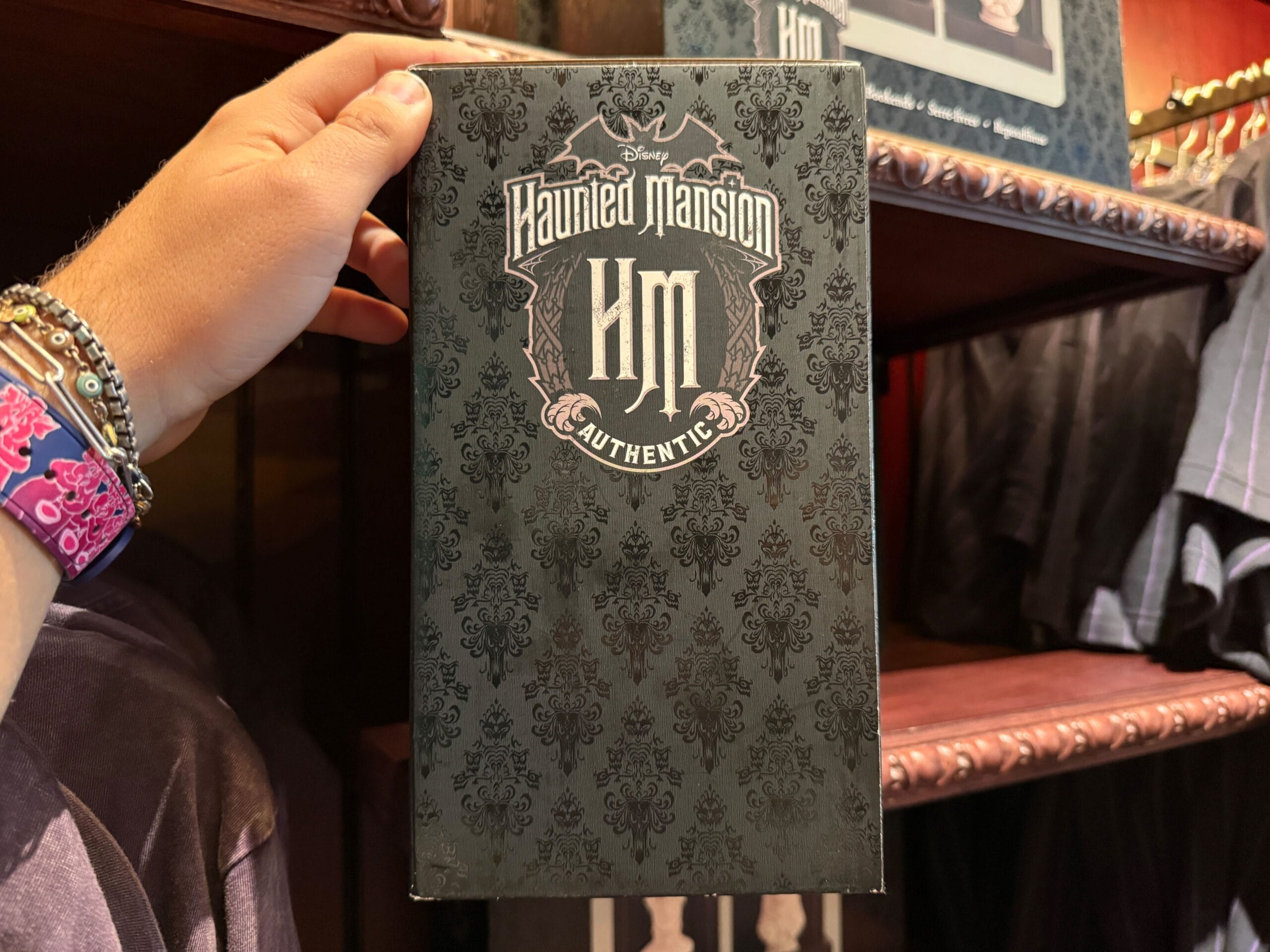 A hand holds Disneyland Haunted Mansion HM Merchandise box with a decorative black pattern in a store.