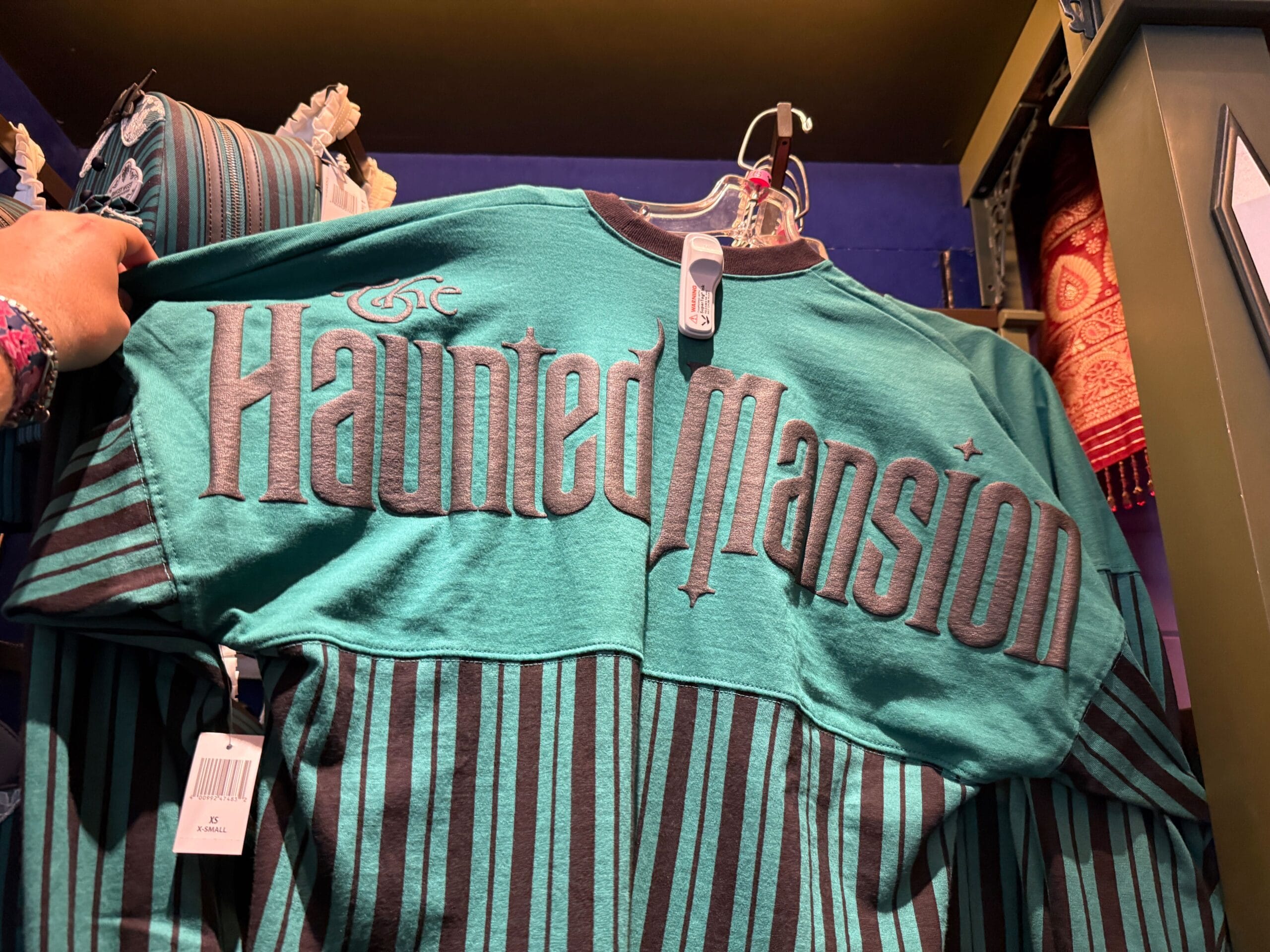 A teal and black striped "Haunted Mansion" shirt, classic HM Merchandise, hangs on a display rack in Disneyland.