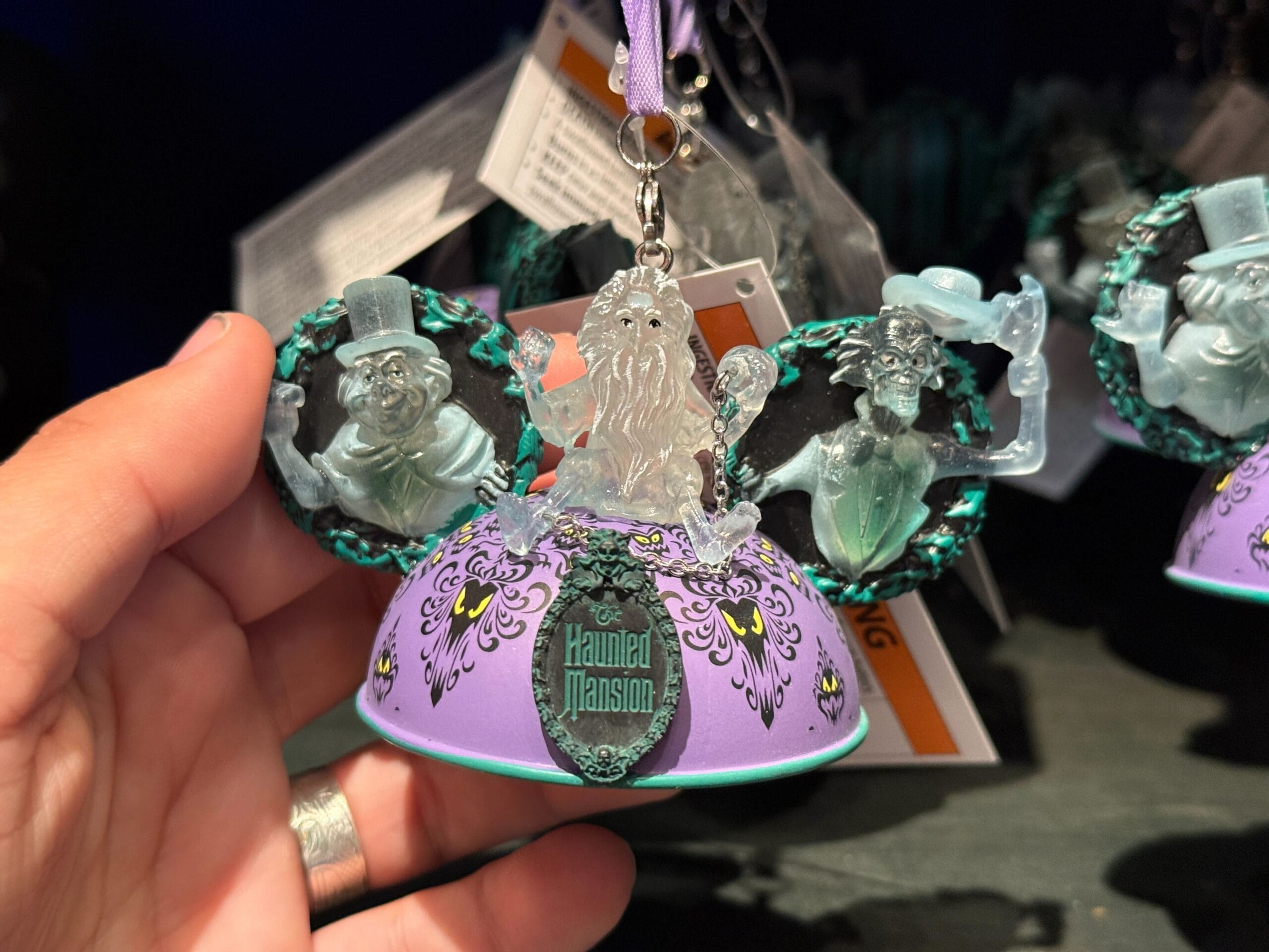 A hand holds a Haunted Mansion ornament from Disneyland HM Merchandise, featuring three ghostly figures on a purple base with Mickey ears.