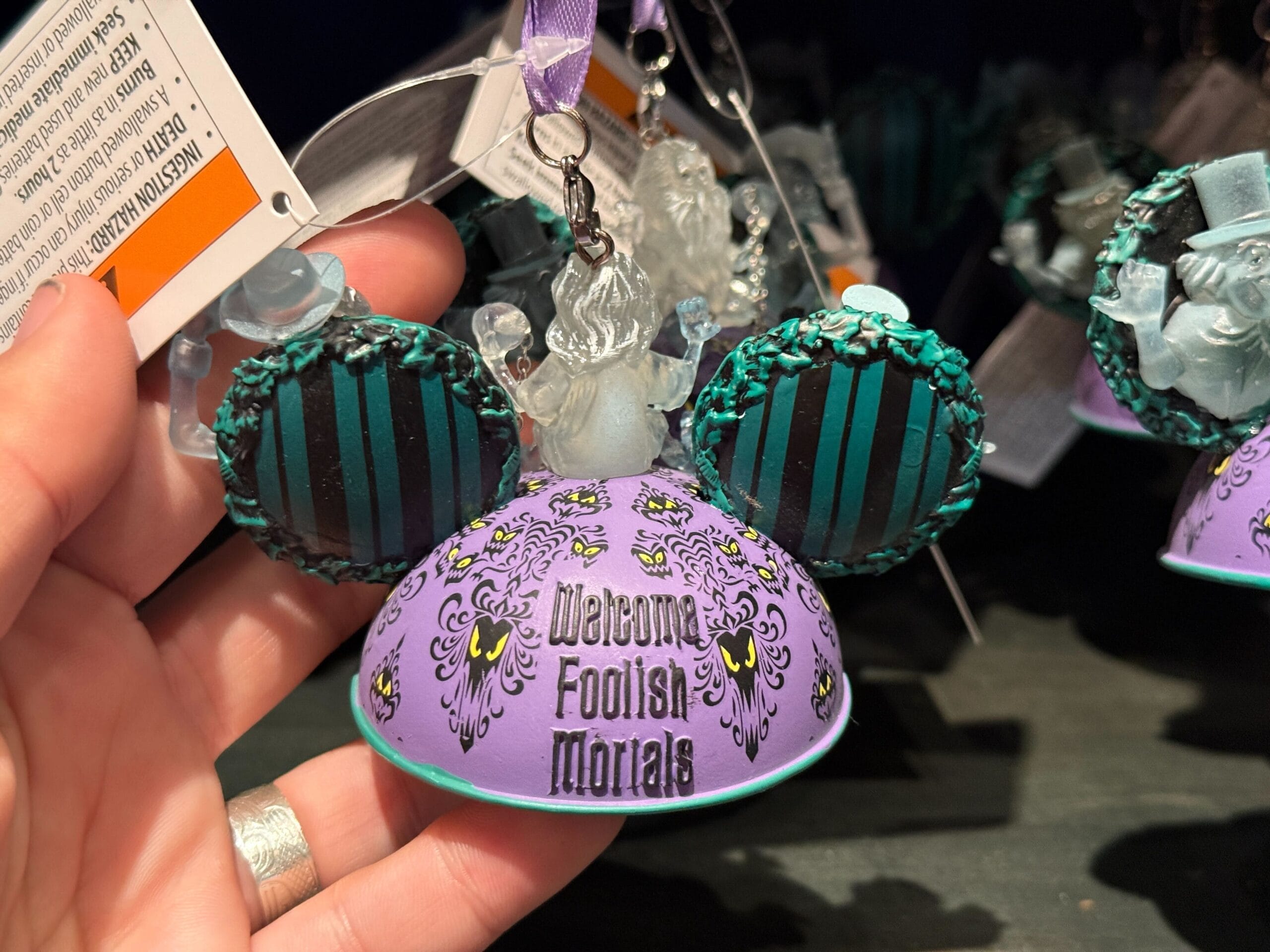 A hand holds a purple "Welcome Foolish Mortals" Mickey ears ornament, a must-have HM Merchandise from Disneyland.