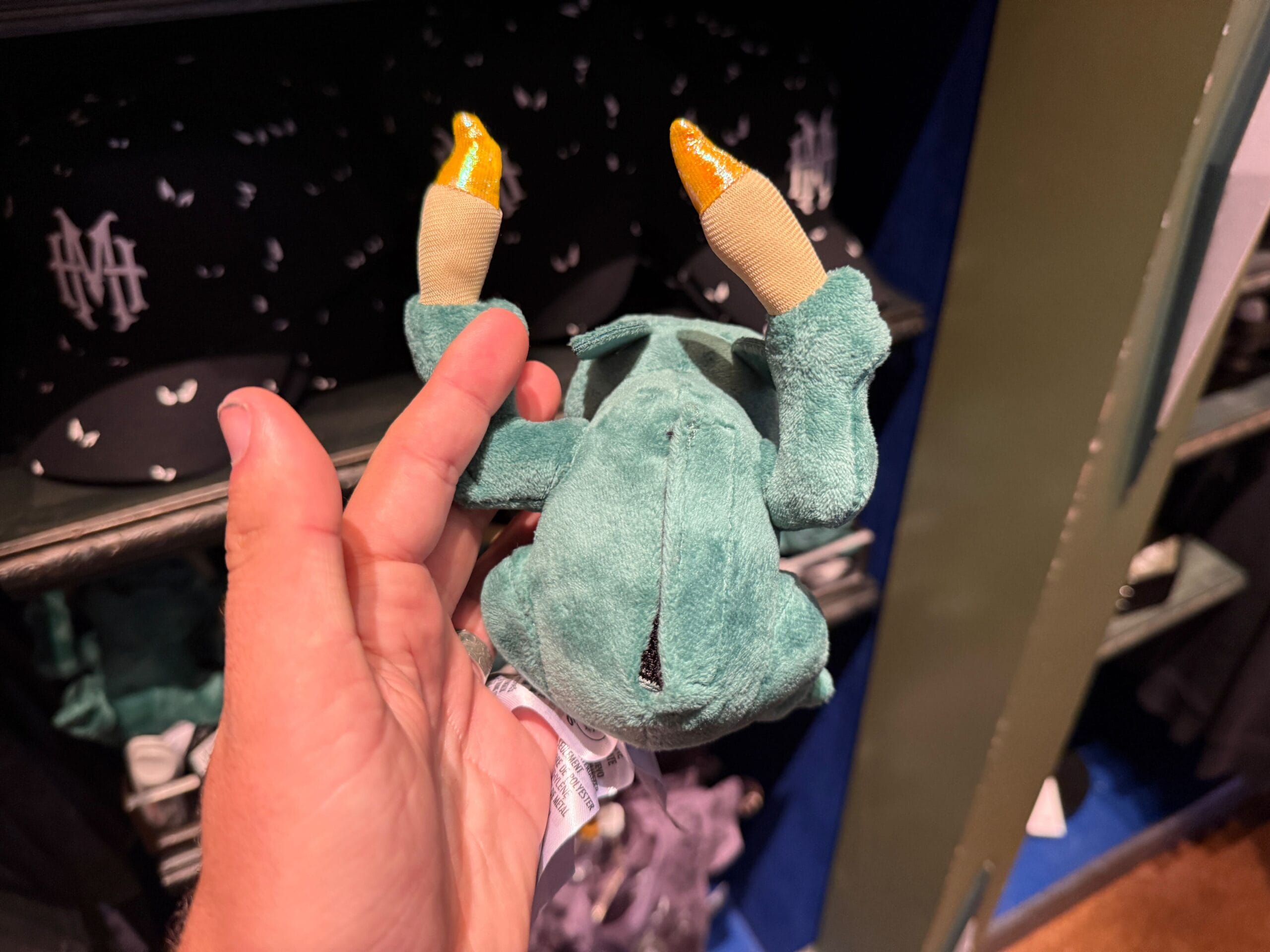 A hand holds a plush toy frog upside down in front of black Disneyland hats with white lettering on a store shelf.